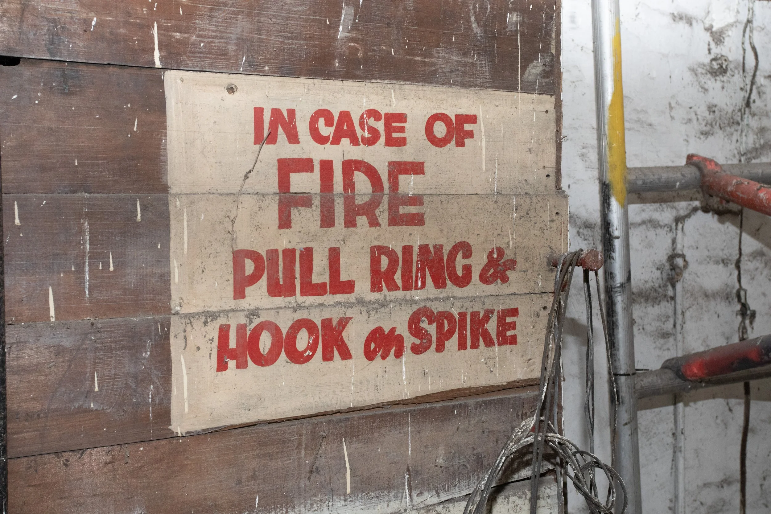 Wooden wall with a sign in red text that reads, 'In case of fire pull ring and hook on spike.' Firefighting equipment in the background.