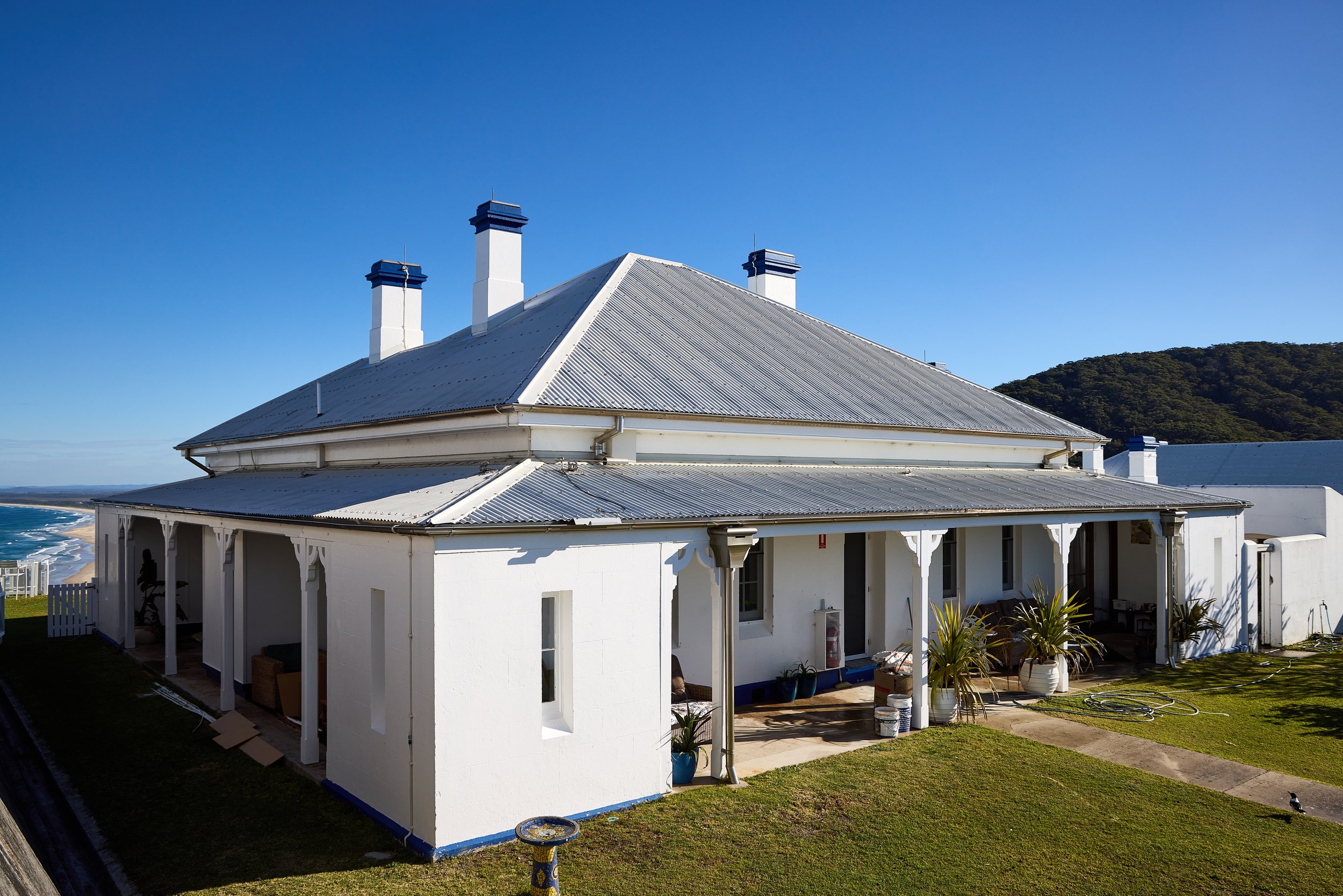 White house with corrugated metal roof and multiple chimneys, surrounded by green lawn and plants, with the ocean and a mountain in the background under a clear blue sky.