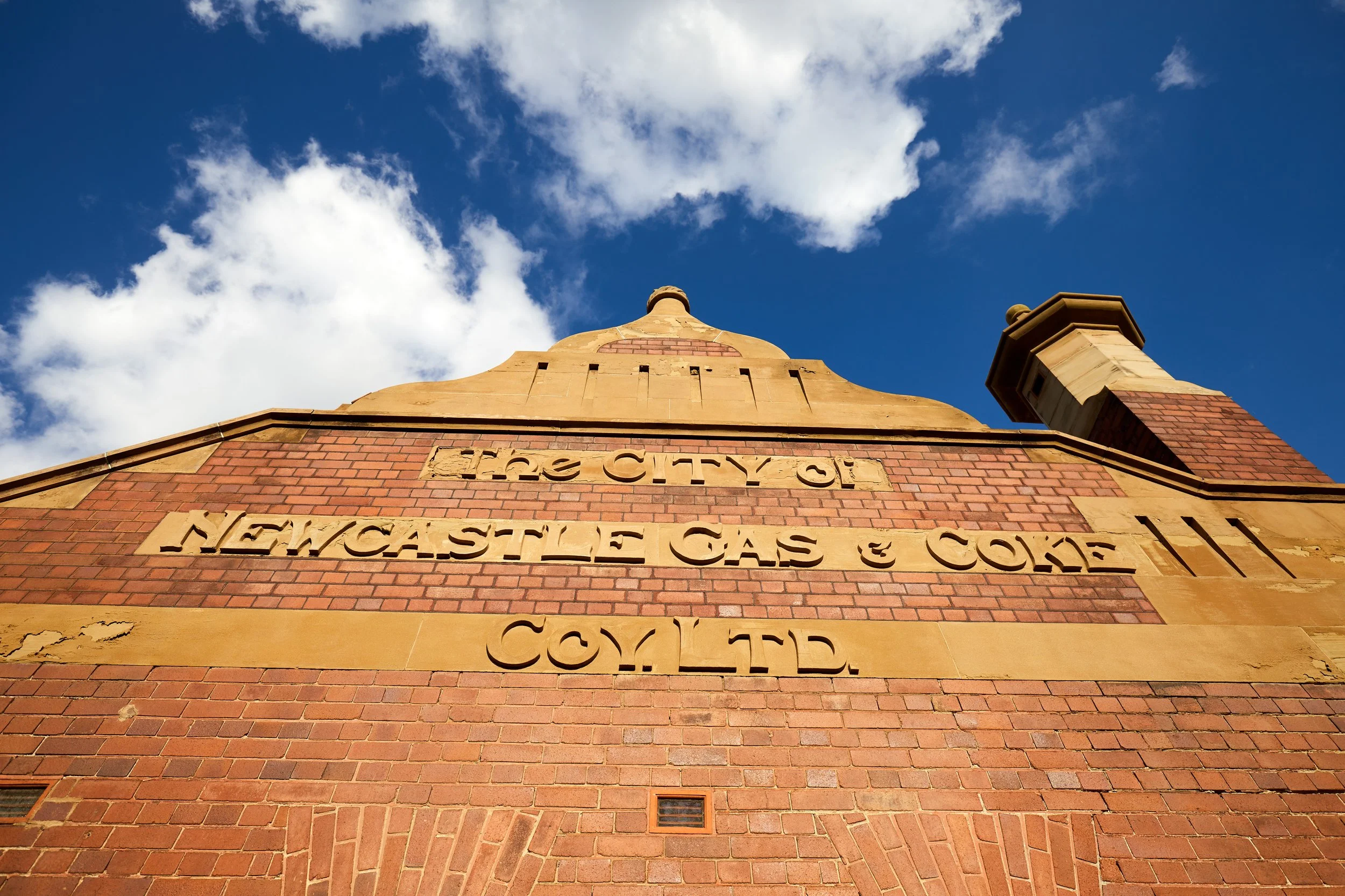 Looking up at a historic brick building with a sandstone dome and chimney, with a bright blue sky and scattered clouds in the background.