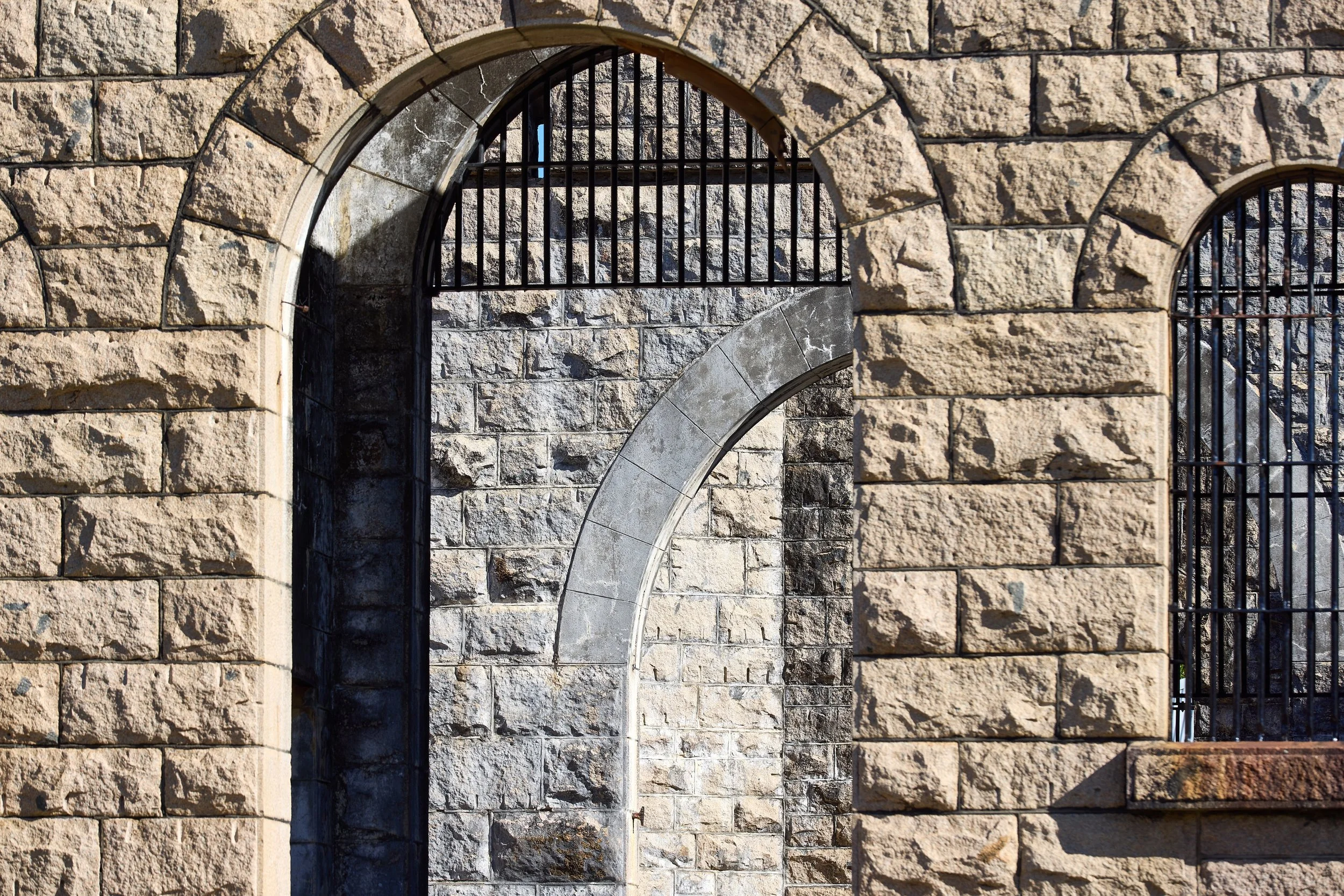 Close-up of stone wall with arched openings and black metal bars, showing weathered stone and shadows.