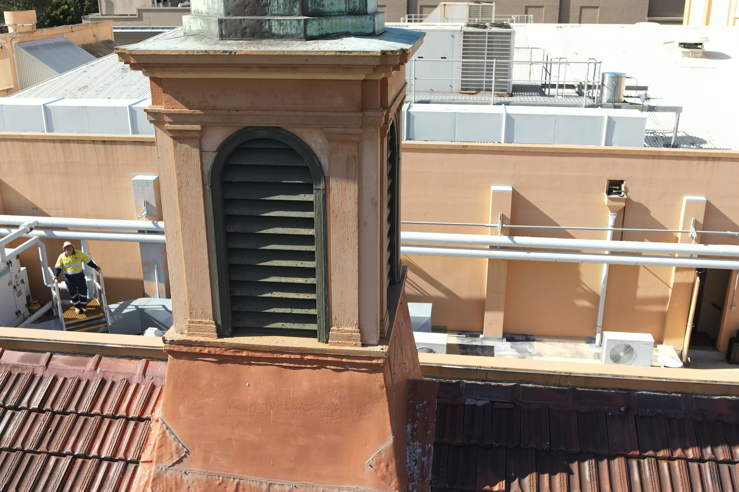 View of a rooftop with various air conditioning units and vents. A worker in a yellow safety jacket and white helmet is on a lift near the bottom left corner.