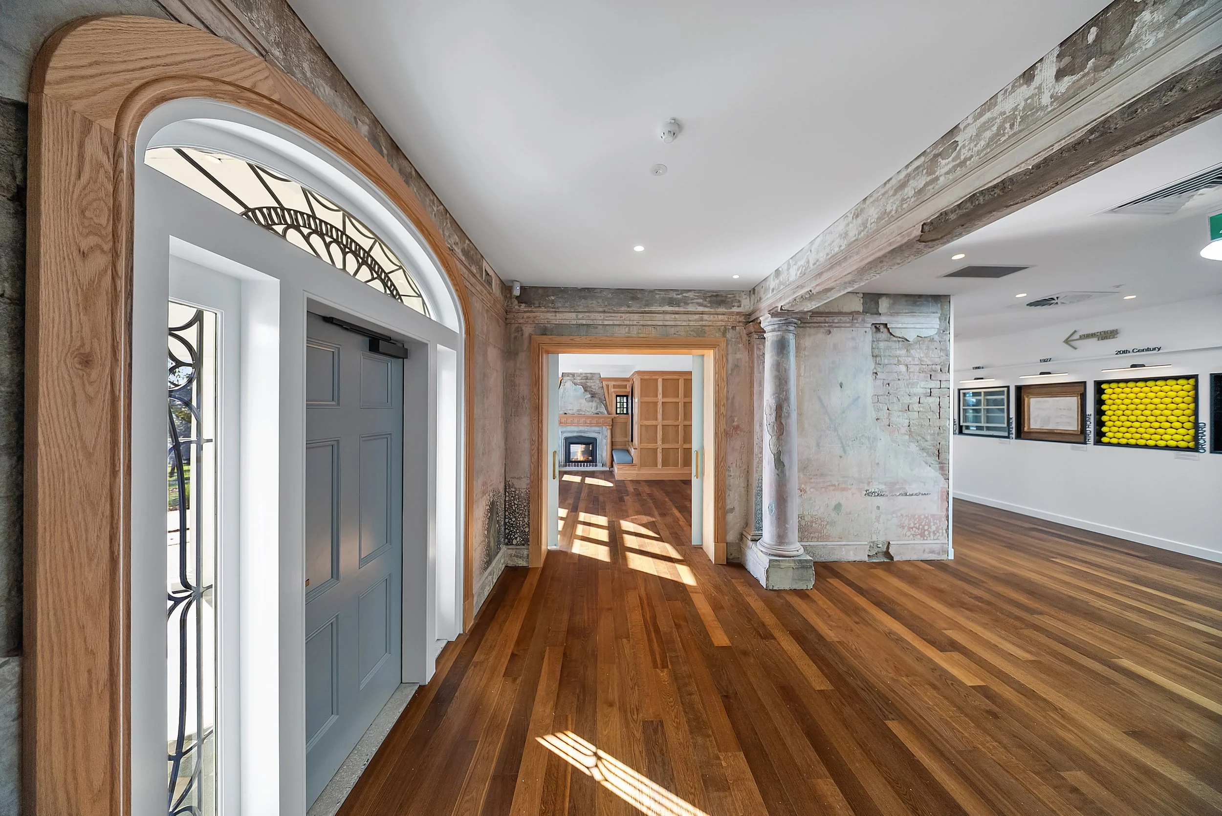Interior of a building with wooden floors, ornate wooden trim around a door, exposed brick and plaster wall areas, and a view through doorways revealing a cozy living room with a fireplace.