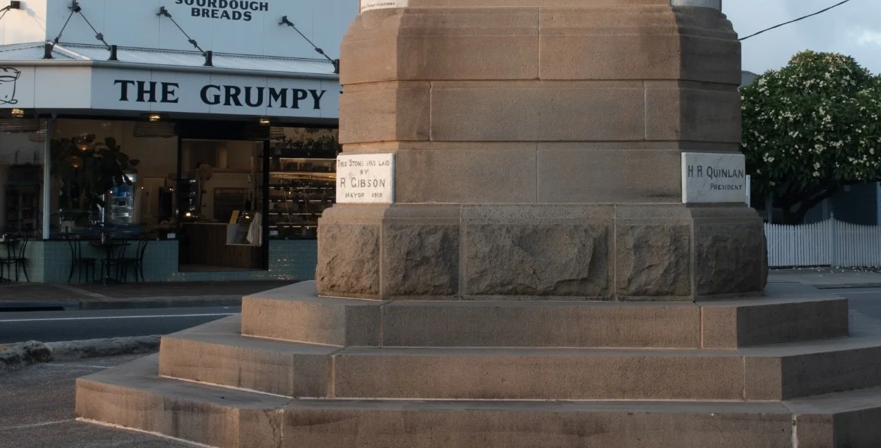 Close-up of a stone monument base with commemorative plaques, in front of a bakery named 'The Grumpy' with a sign advertising sourdough bread. There is a tree with white flowers and a white picket fence in the background.