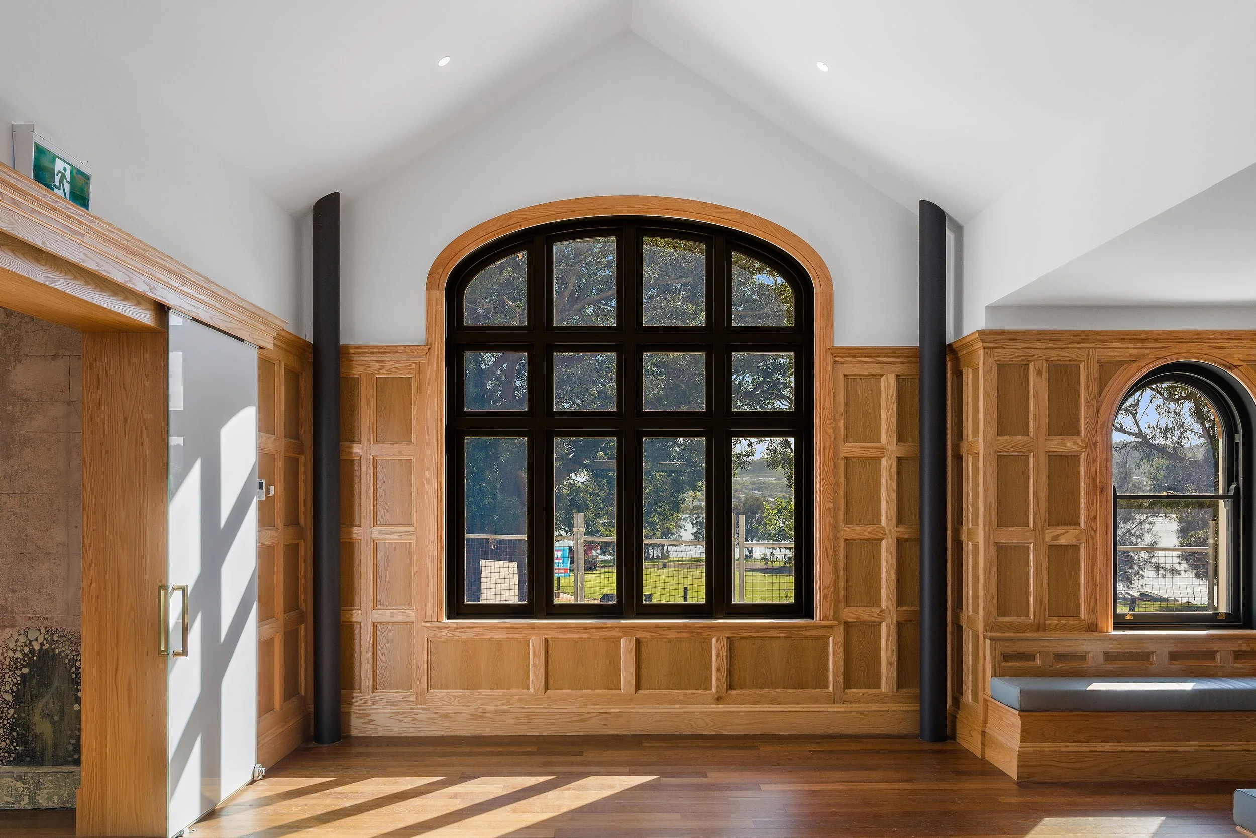 Interior of a modern house with large arched window, wooden wall paneling, and built-in seating, showing trees and a street view outside.