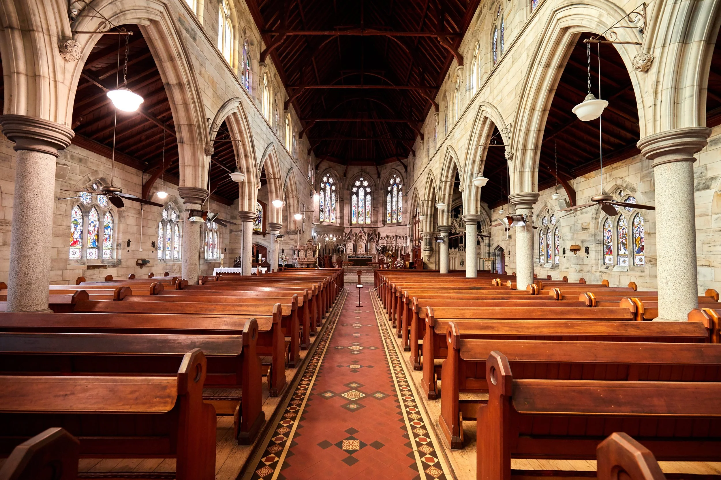 Interior of a church with wooden pews, stained glass windows, stone arches, and hanging ceiling fans.