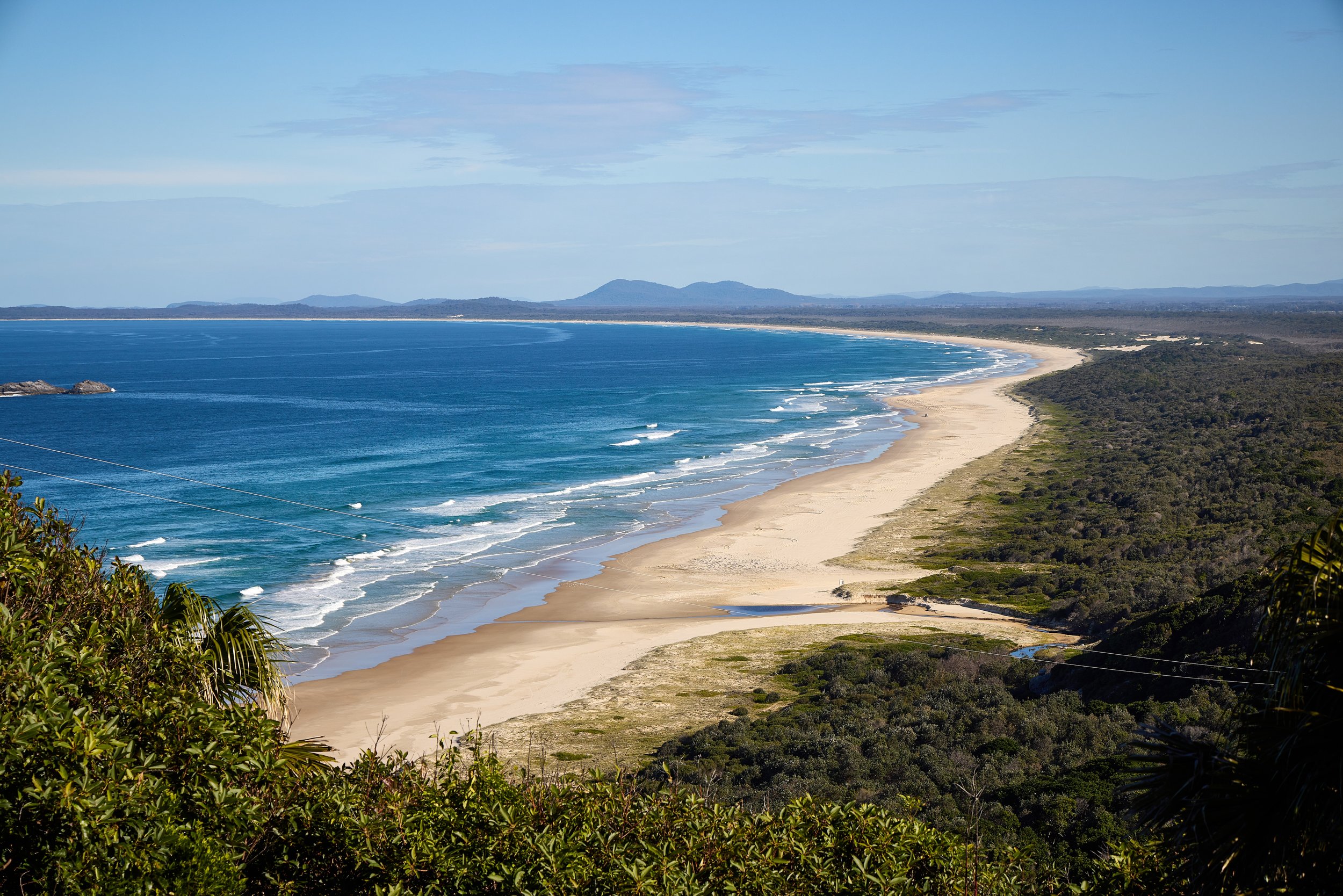 A scenic view of a sandy beach along the coastline with waves gently hitting the shore, bordered by lush green vegetation and distant hills under a partly cloudy sky.