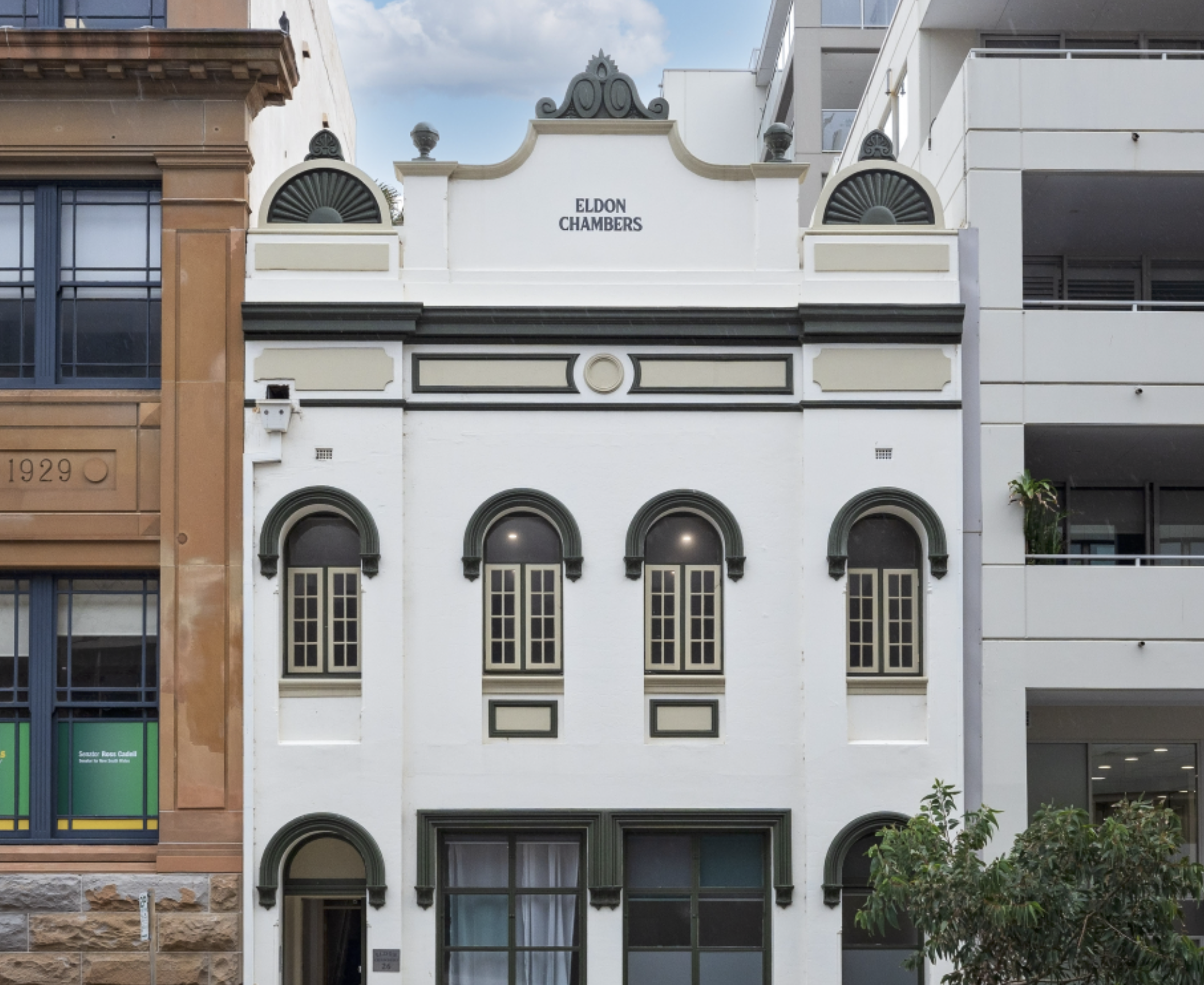 The image shows the front facade of a building with the name 'Eldon Chambers' displayed at the top center. It features decorative architectural elements, large windows, arched doorways, and a mix of white, beige, and dark green colors. The building is flanked by other buildings, one brown on the left and a white modern-looking building on the right. There is a small tree in front of the building.
