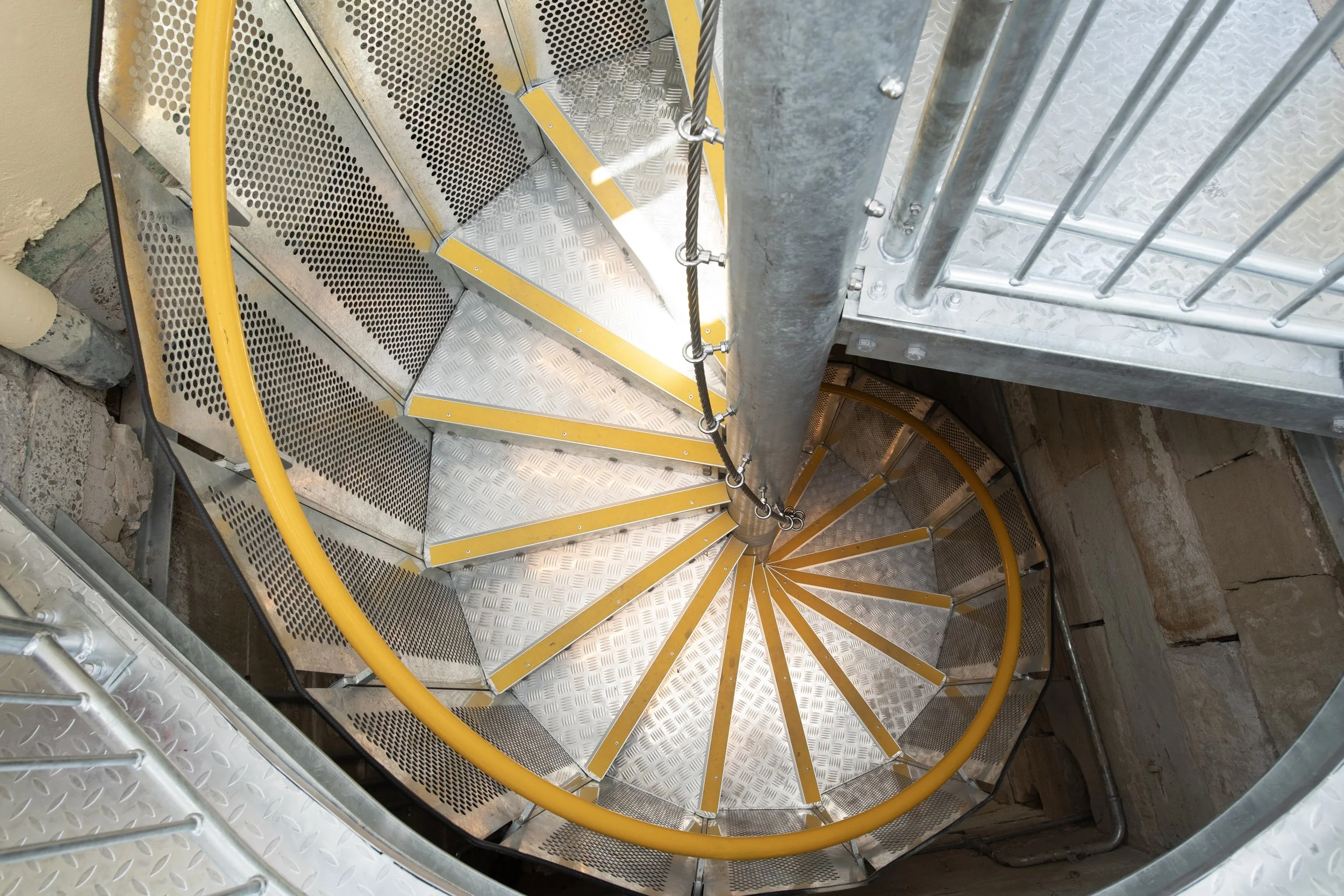 Top-view of a spiral staircase made of metal, with yellow handrails and diamond-patterned steps, surrounded by concrete and metal rails.