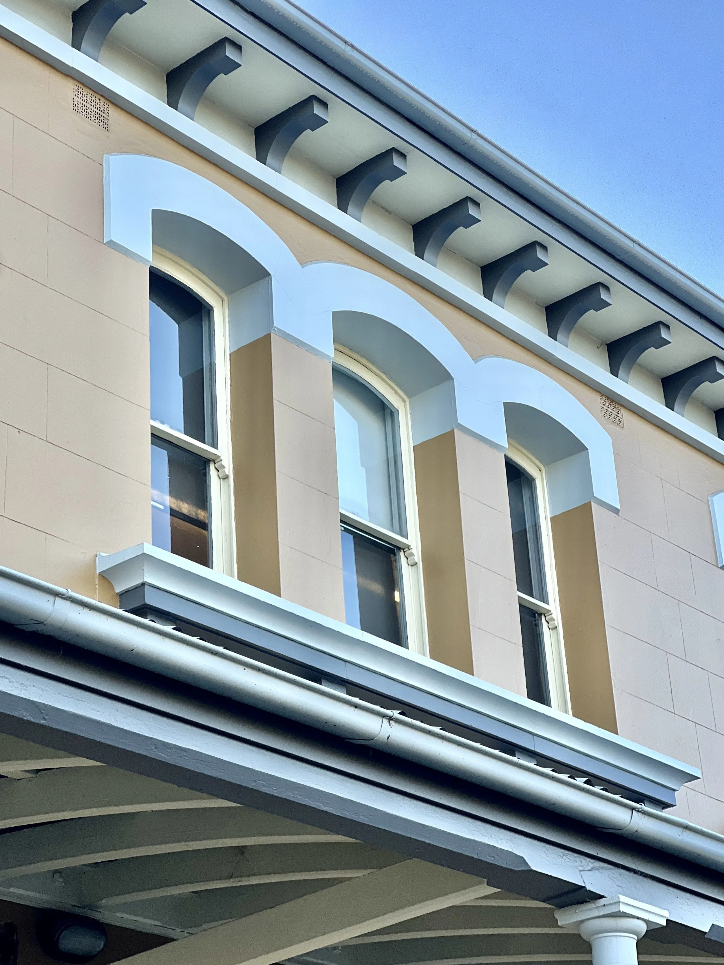 Close-up photo of the upper part of a beige building with arched windows, decorative molding, and balcony supports, under a clear blue sky.