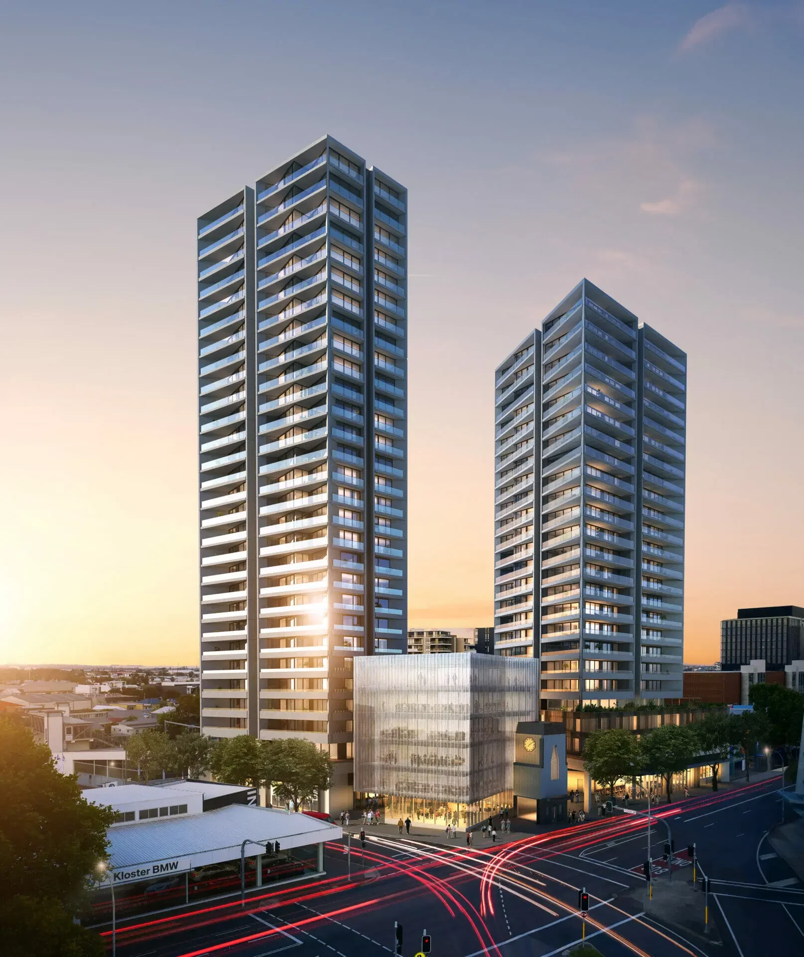 Two modern high-rise apartment buildings at sunset in an urban area, with a busy intersection and light trails from moving vehicles in the foreground.
