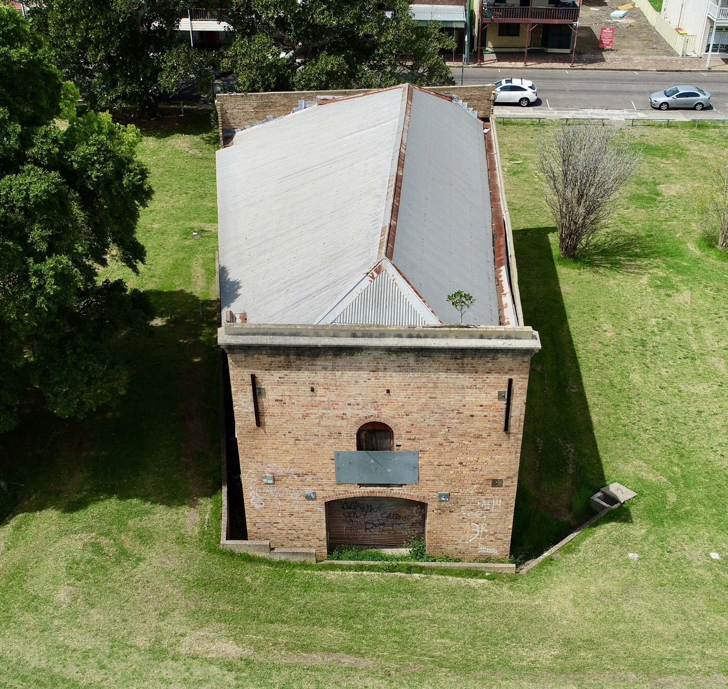 Aerial view of a brick building with a white metal roof, surrounded by green grass, trees, and parked cars on a nearby street.