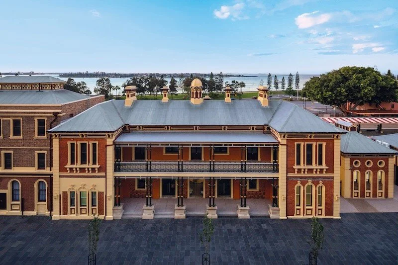 Historical brick building with a covered porch and decorative architectural details, overlooking a waterfront with trees and a cloudy sky.