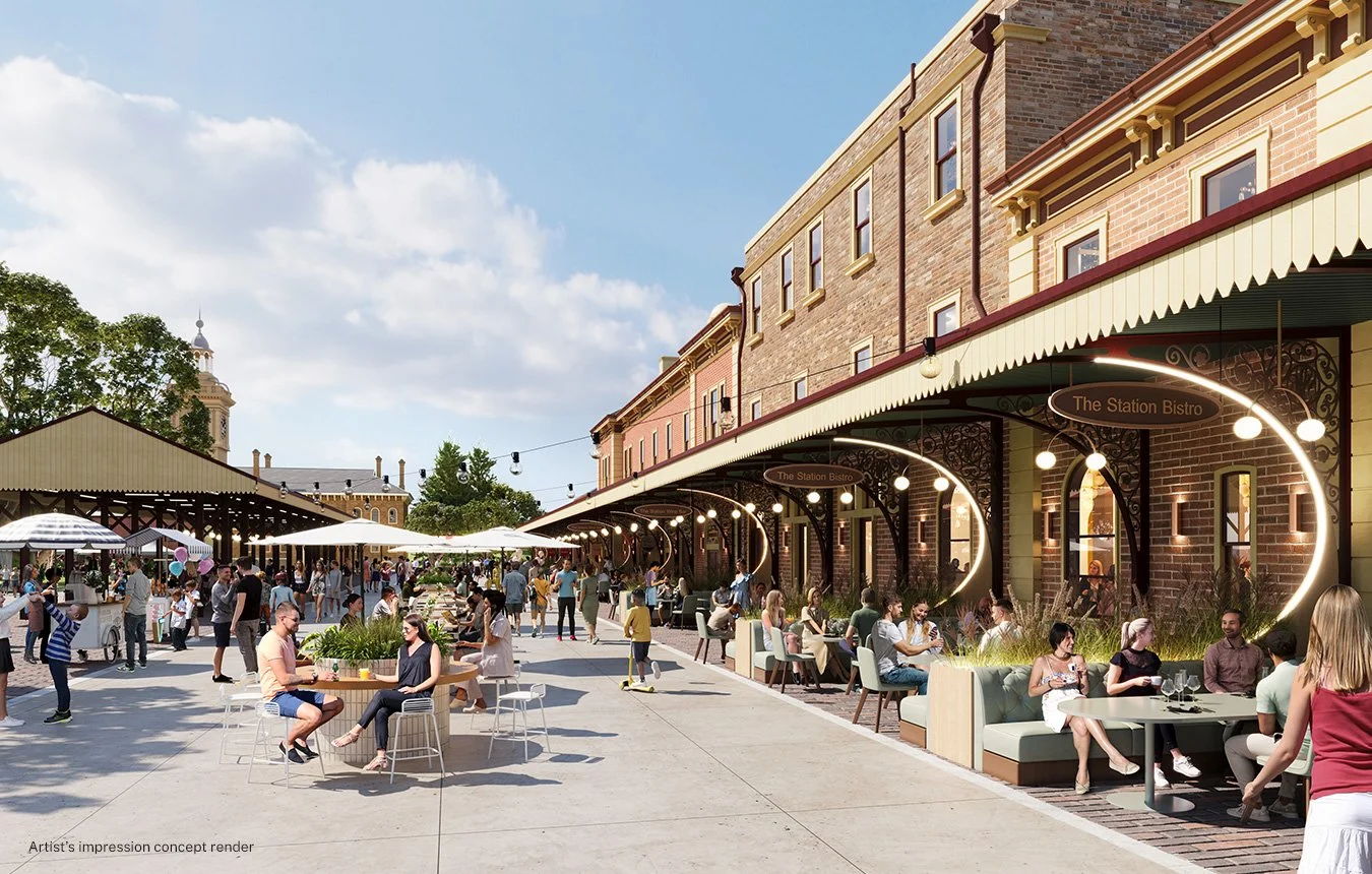 An outdoor marketplace with people dining at a bistro called The Station Bistro, featuring illuminated circular signs and brick buildings, under a blue sky with clouds.