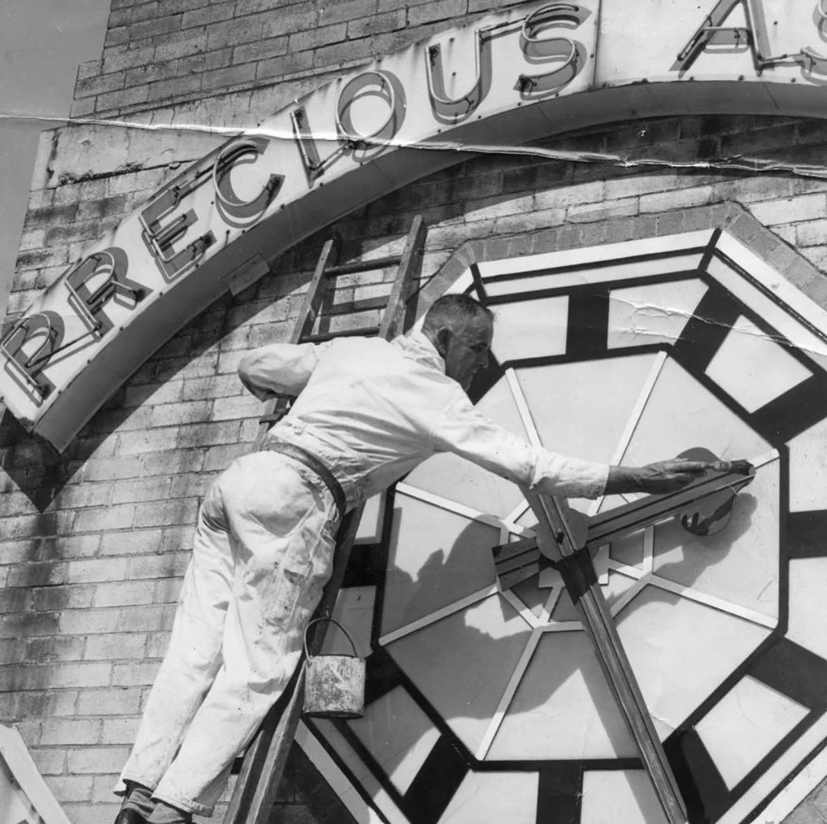 A man painting a large clock face on a brick building, with a sign above that reads 'PERFECT CLOCK'.