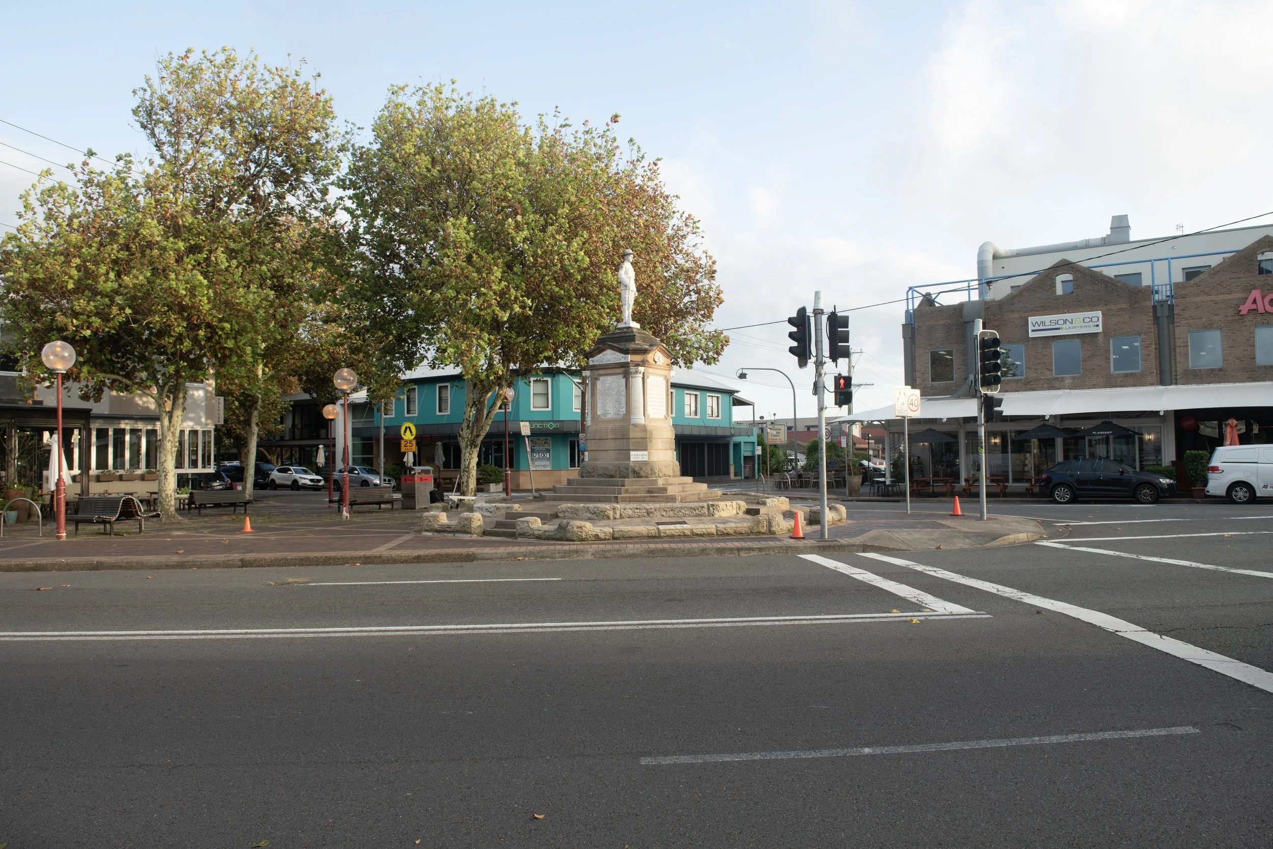 A small town square with a monument in the center surrounded by trees, benches, streetlights, cars, and shops. Traffic lights and crosswalks are visible.