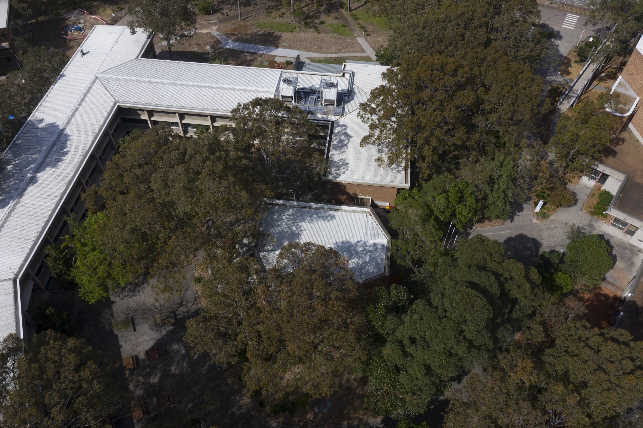 An aerial view of a college or university campus with multiple buildings, trees, and pathways. The roofs of the buildings are mostly white, and there are parking spaces and a small section of road visible.