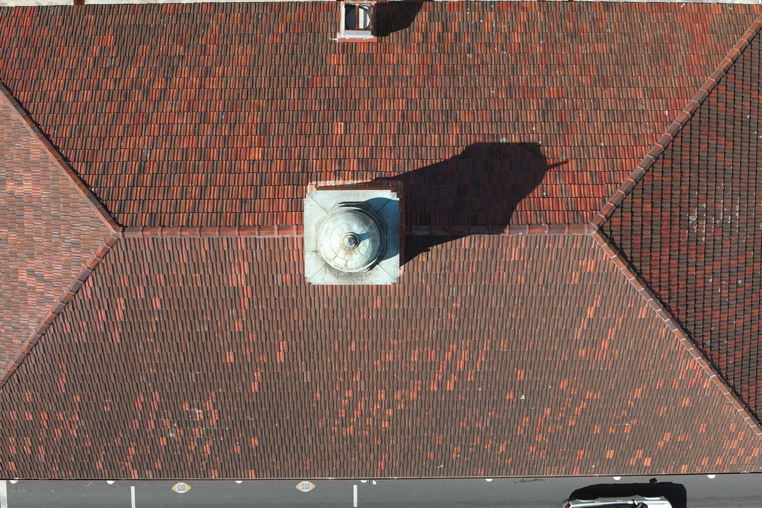 Aerial view of a roof with red clay tiles, including a chimney and shadow of a person taking the photo. The roof has a triangular shape with ridges separating sections, and a small vent near the top.