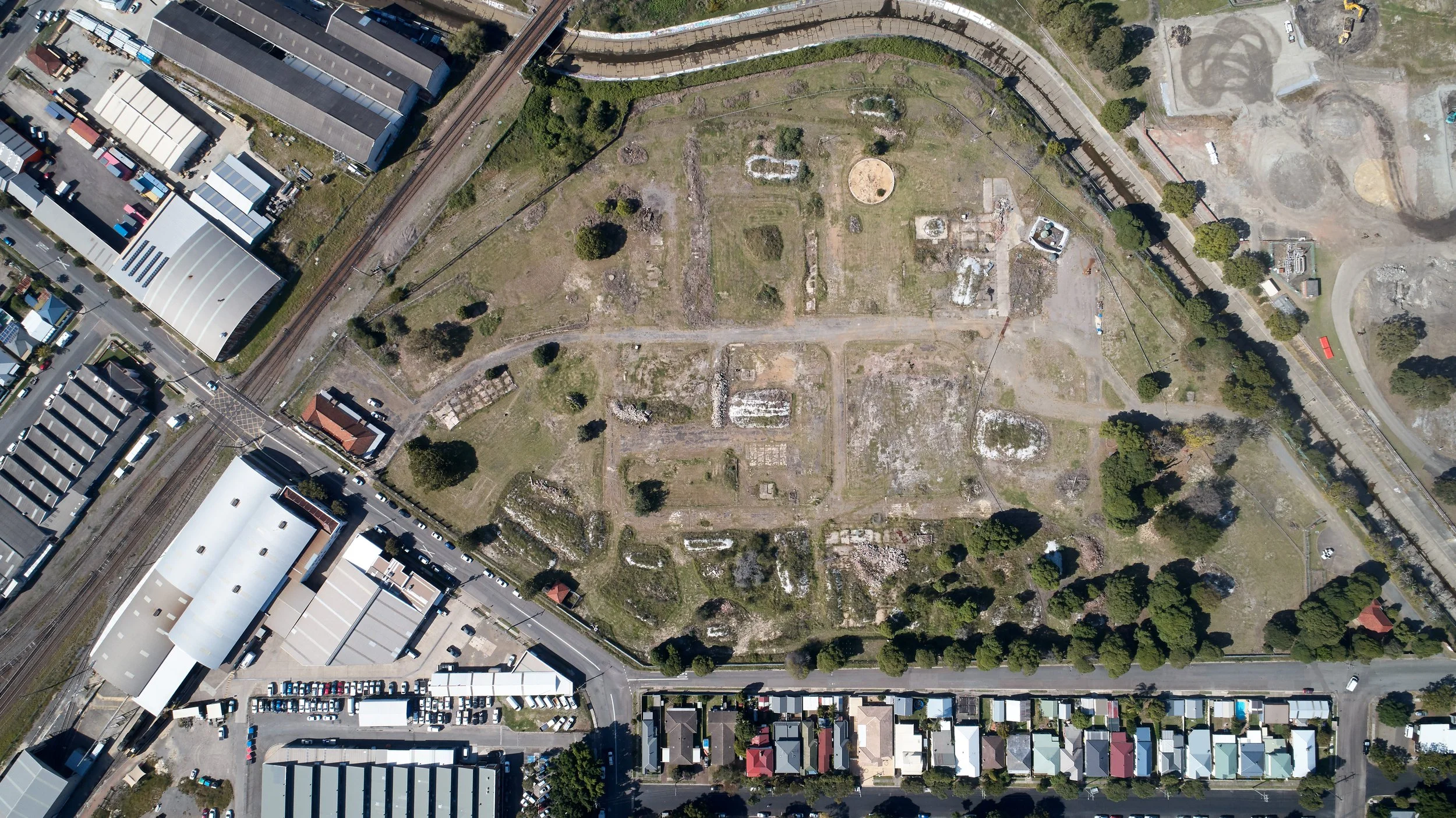 Aerial view of an archaeological site with ruins and green spaces, surrounded by buildings, parking lots, and train tracks.
