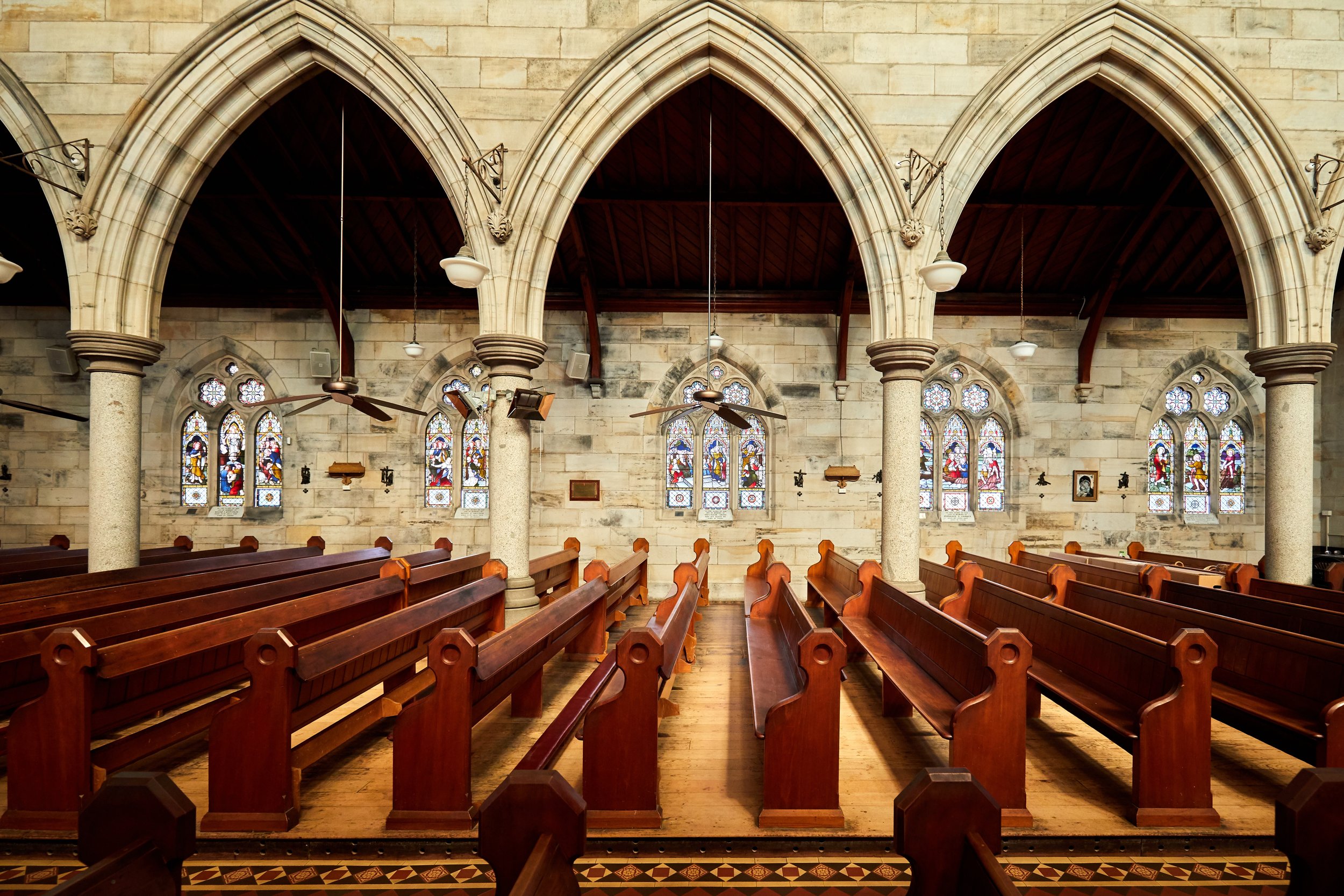 Interior of a church with wooden pews, stained glass windows, stone arches, hanging light fixtures, and ceiling fans.