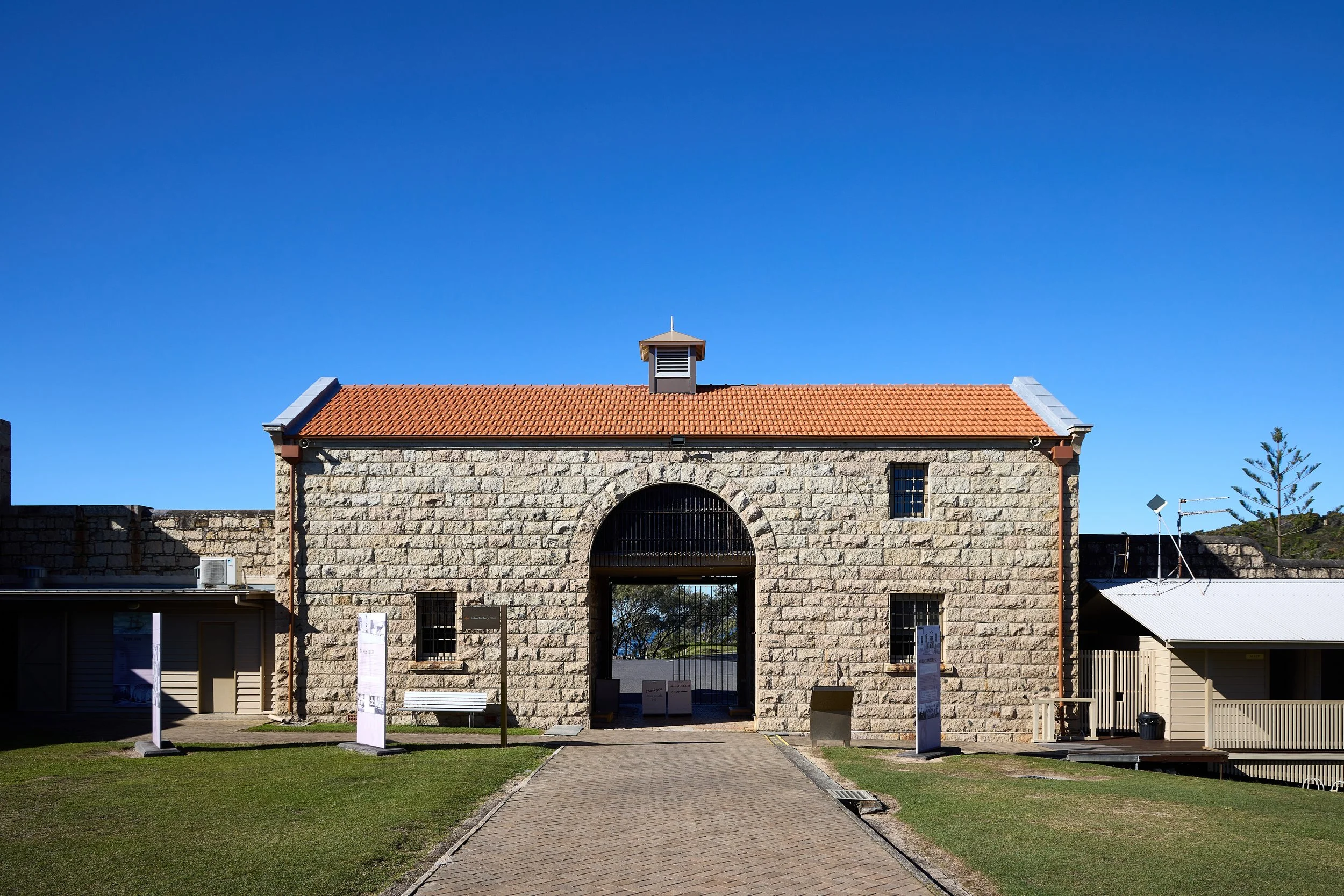 A stone building with a red-tiled roof, small windows, a large archway entrance, and a smaller roofed tower at the center of the roof, set against a clear blue sky. There is a paved pathway leading to the entrance, and information boards and signs ar