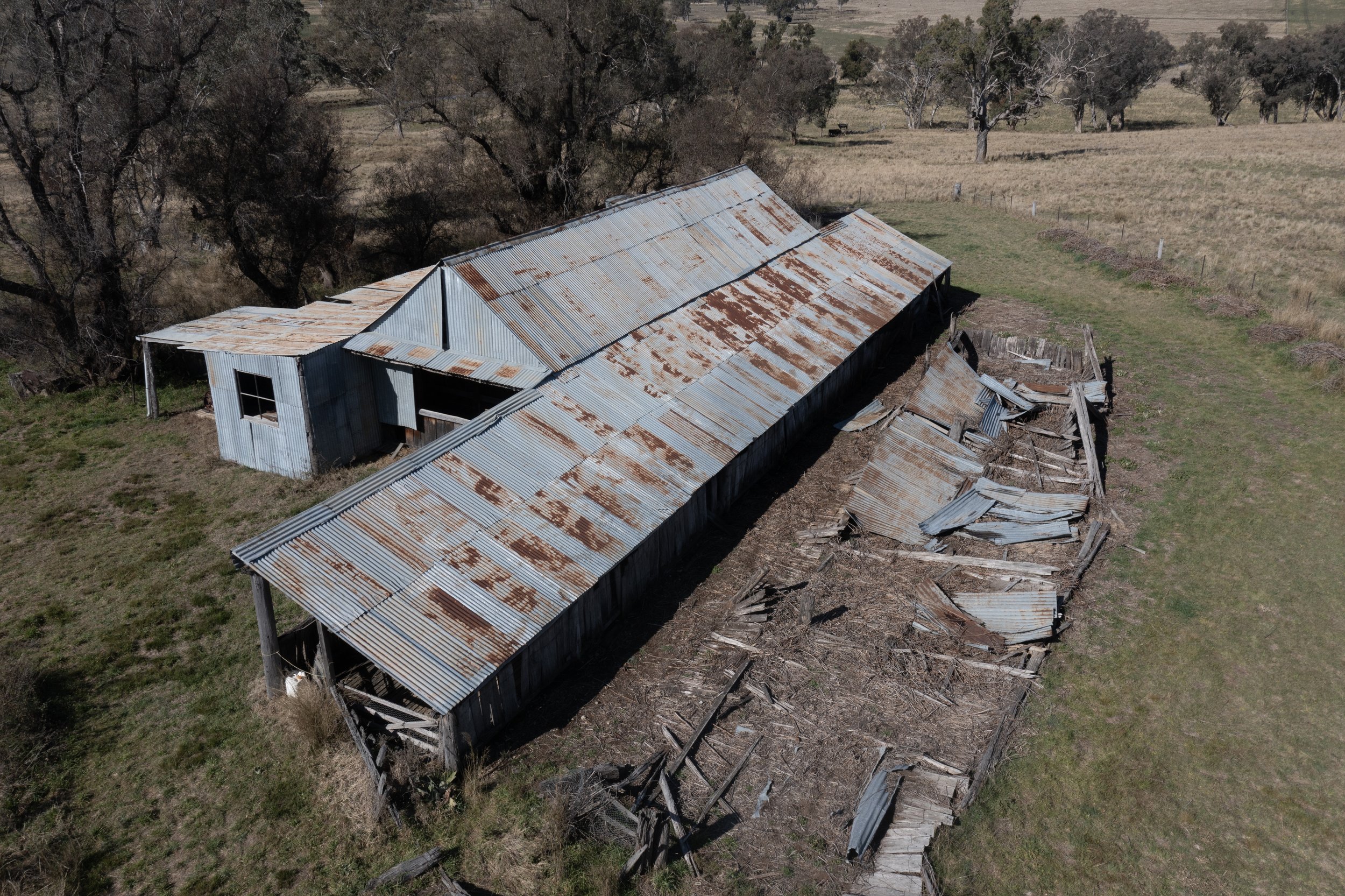 An aerial view of an abandoned, dilapidated barn with rusted corrugated metal roof situated in a rural landscape with sparse trees and open fields.