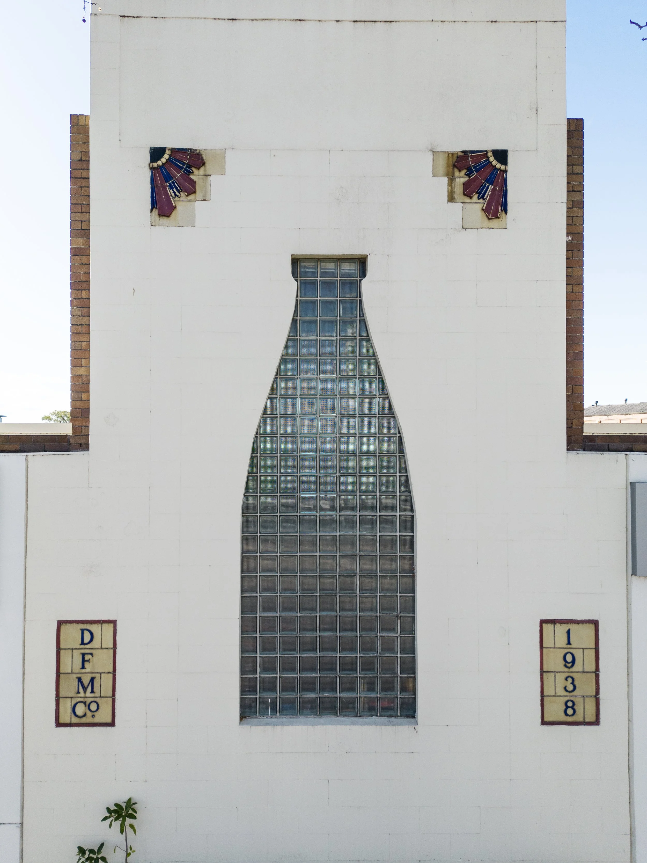 A white building with a tall, narrow stained glass window in the center. Two small decorative tiles with colorful fan designs are near the top. The sky is blue, and there are small plants near the base of the building.