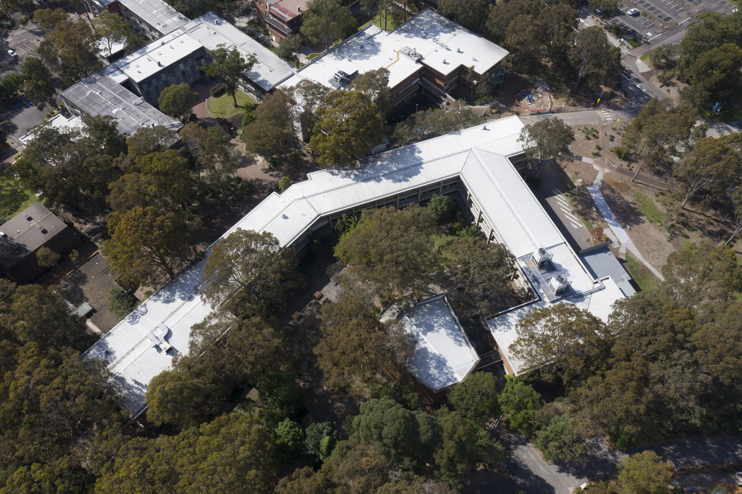 Aerial view of a university campus with multiple white-roofed buildings surrounded by trees and roads, with parking lots visible to the side.