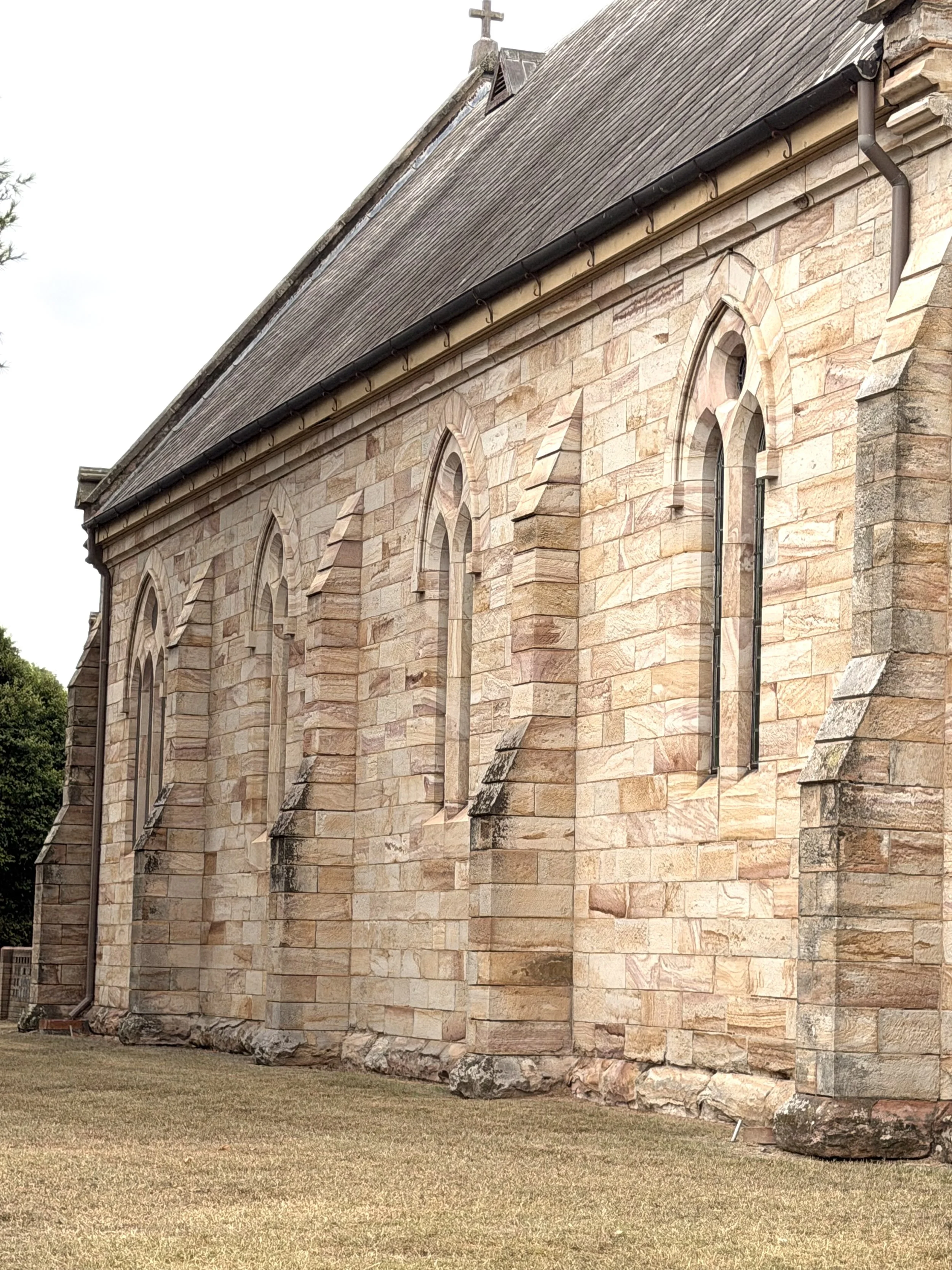 Side view of an old stone church with pointed arch windows, a cross on the roof, and a well-maintained lawn in front.