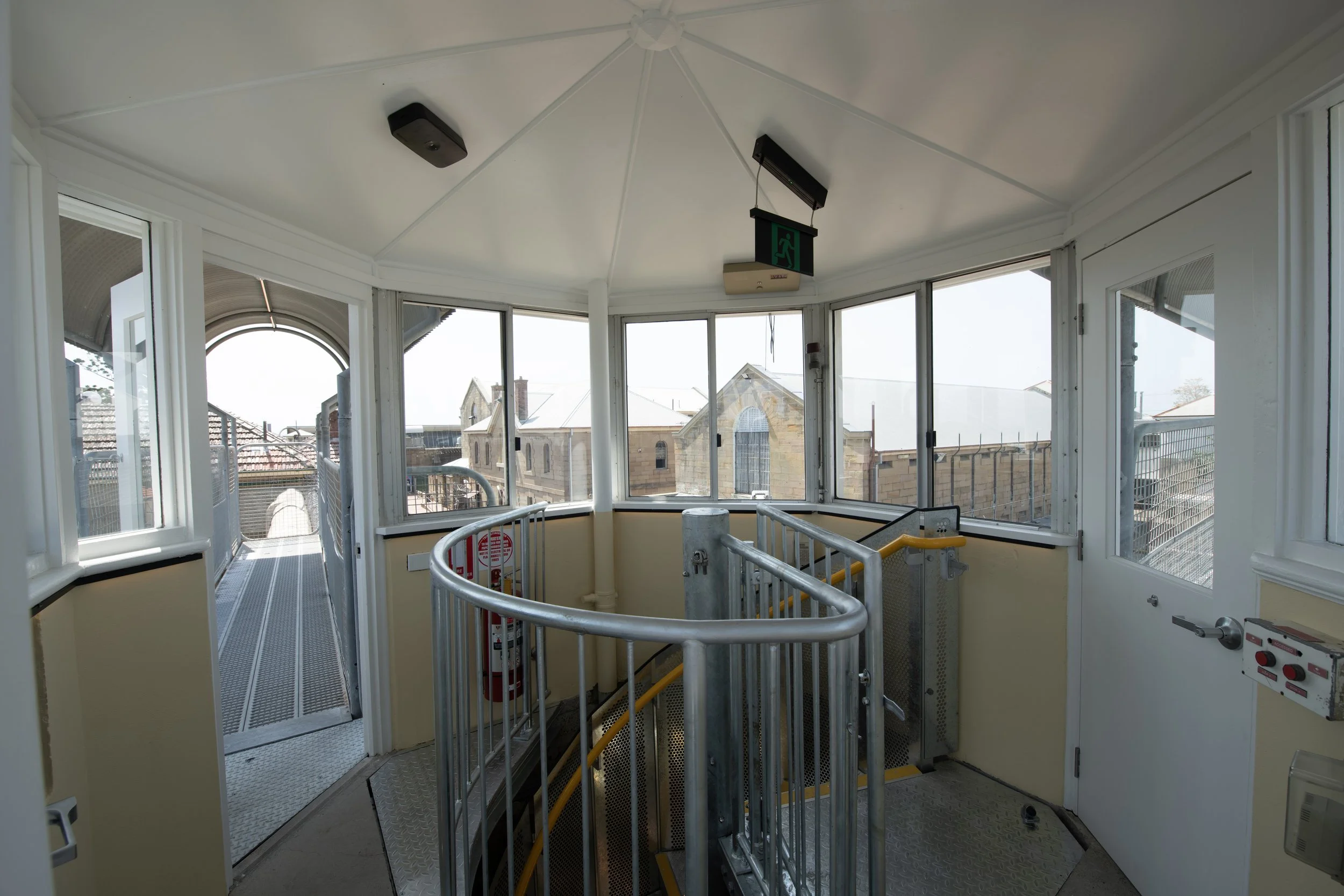Interior of an elevator with large windows, view of buildings outside, safety signs, and a staircase at the bottom.