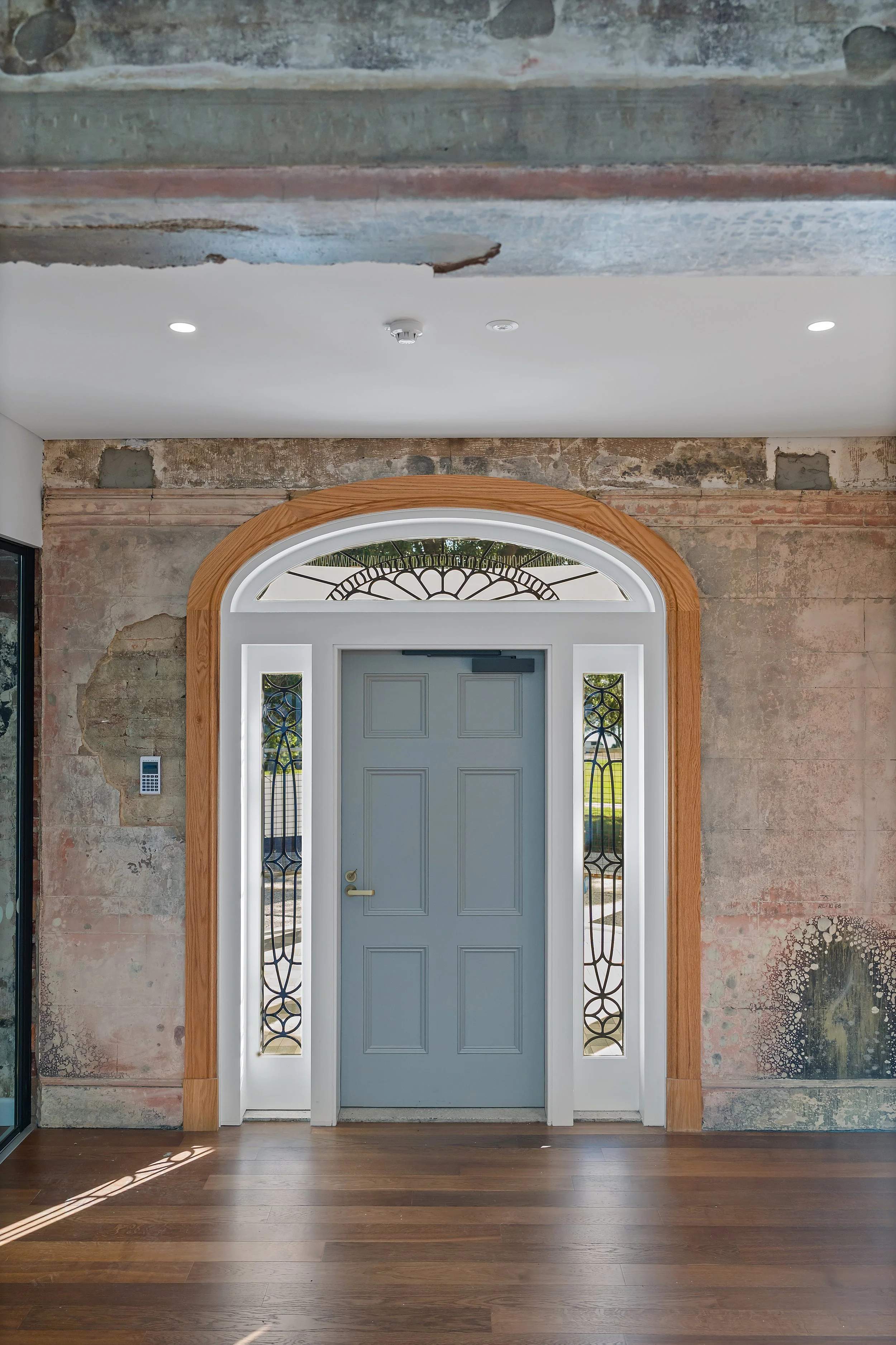 Front door painted blue with sidelights featuring decorative wrought iron designs, set in an archway with wood trim, surrounded by a partially renovated exposed brick wall and wood flooring.
