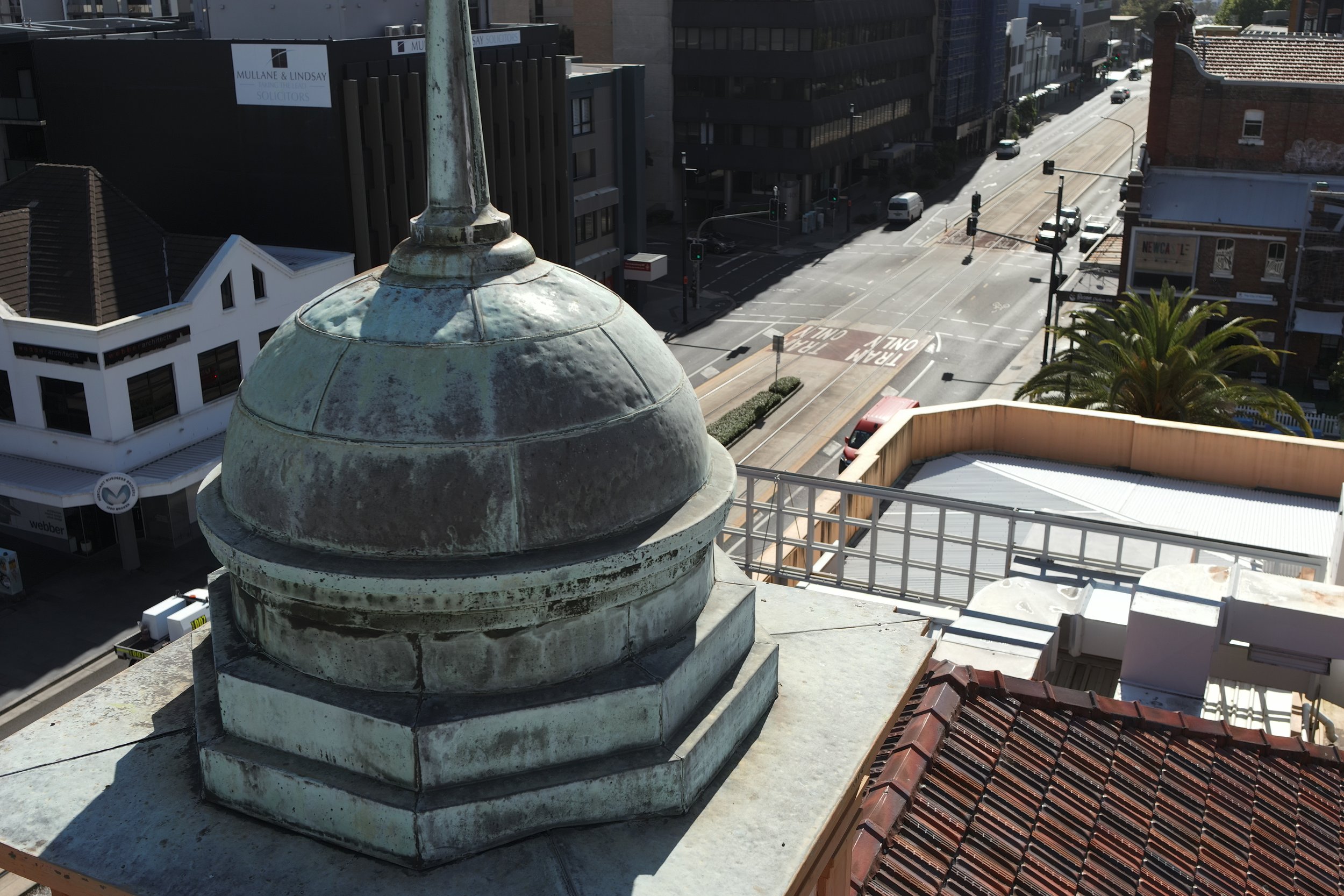 View from a high rooftop showing an old, weathered dome-shaped chimney, city streets with cars, traffic lights, and surrounding buildings including a modern dark building, a white building with signs, and a street with tram tracks.