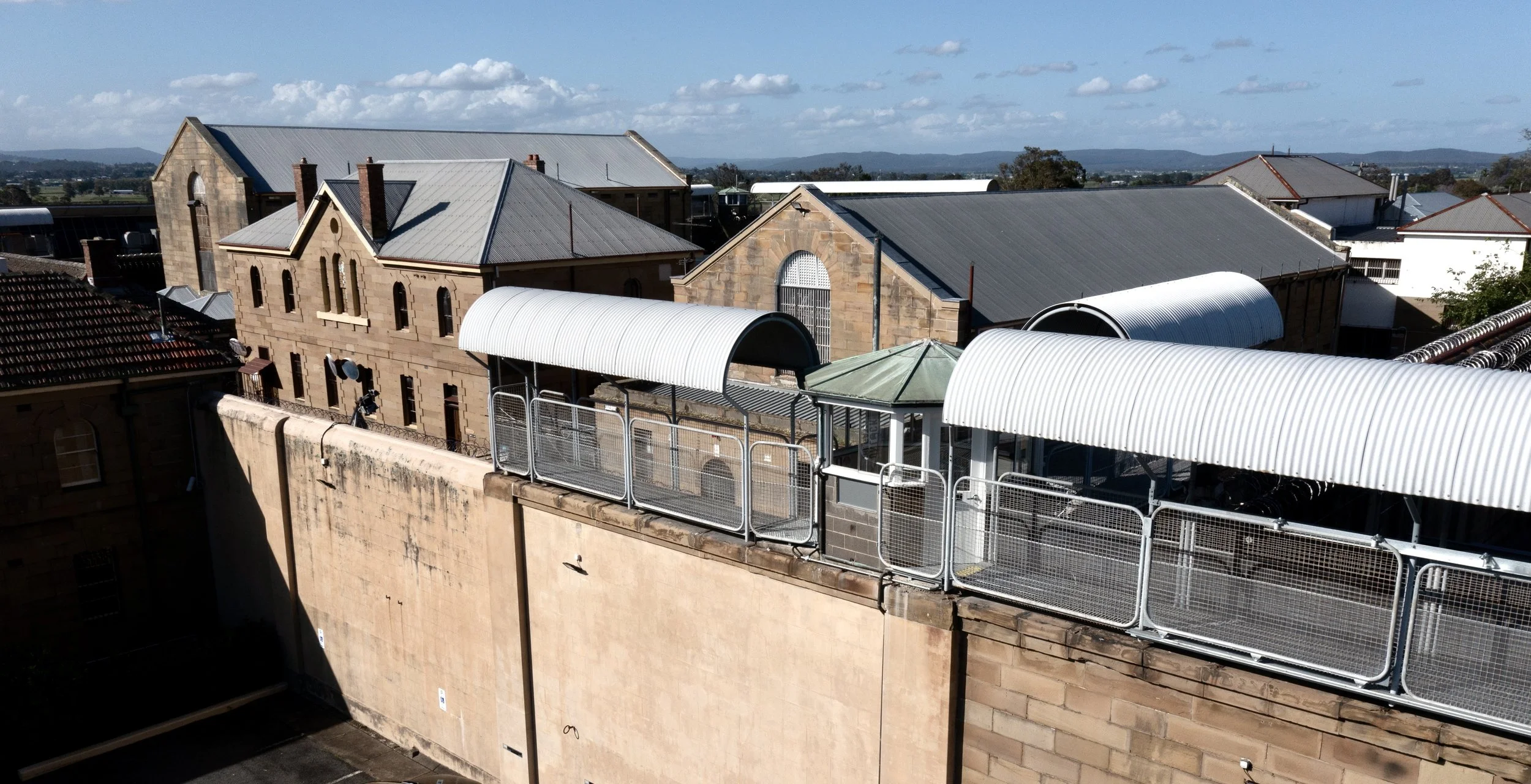 View of rooftops with brick and stone buildings, metal roofs, and a rooftop terrace with white-striped awnings and fencing, under a partly cloudy sky.