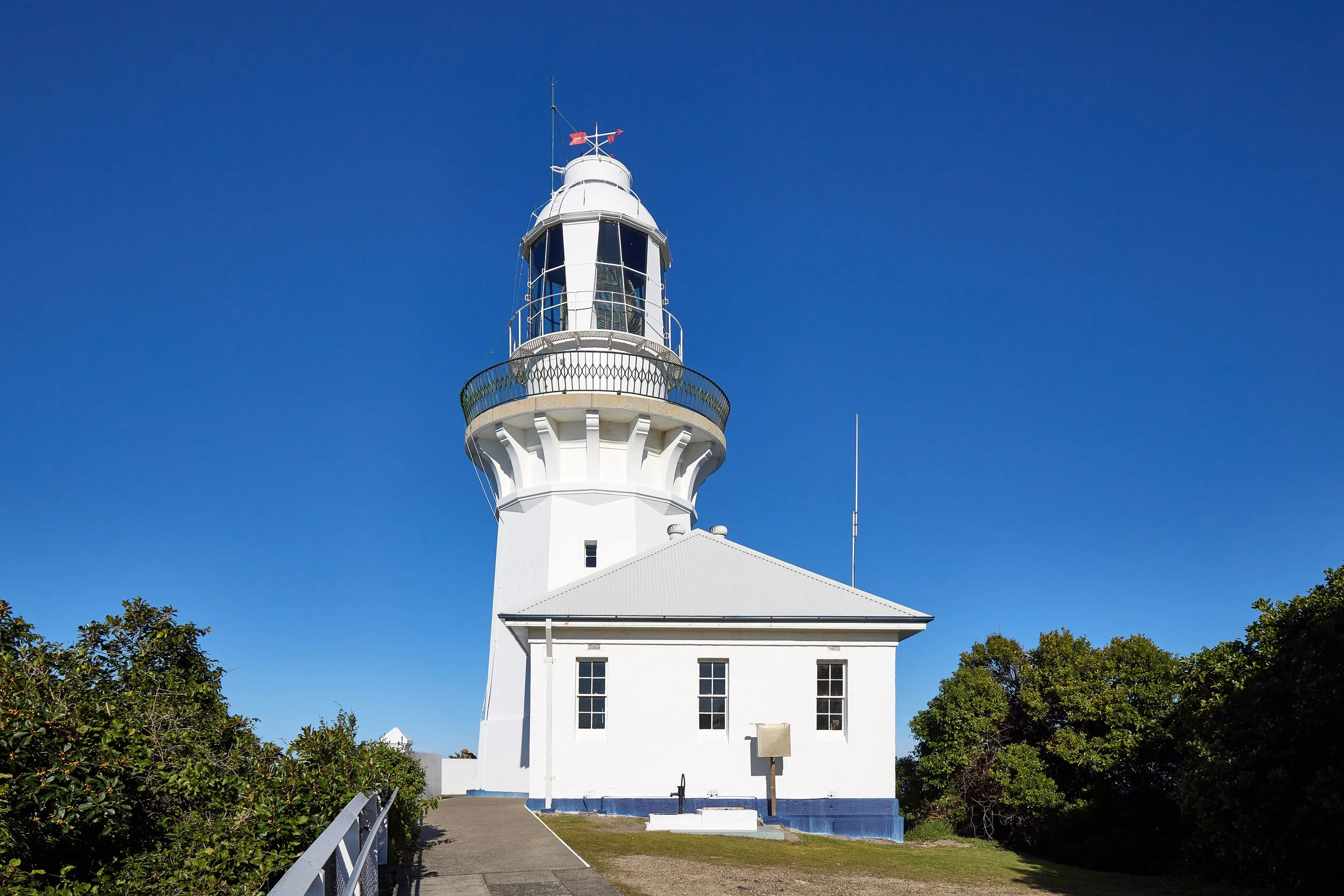 A white lighthouse with a black lantern room on top, set against a clear blue sky, surrounded by green trees and a small pathway leading up to it.