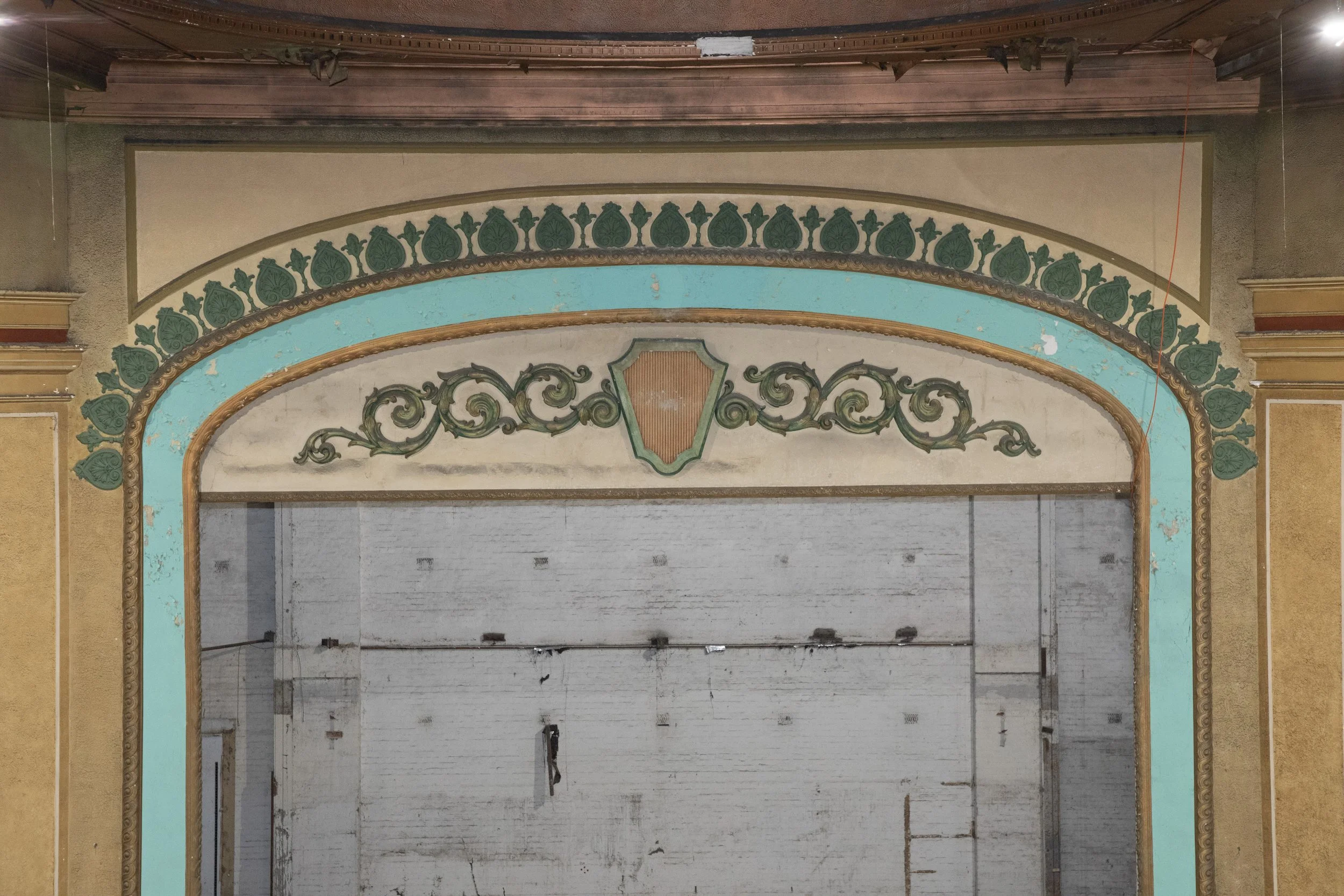 Photograph of an old, ornate theater or auditorium stage with decorative trim, floral pattern, and a partially visible proscenium arch. The stage appears empty and somewhat worn.