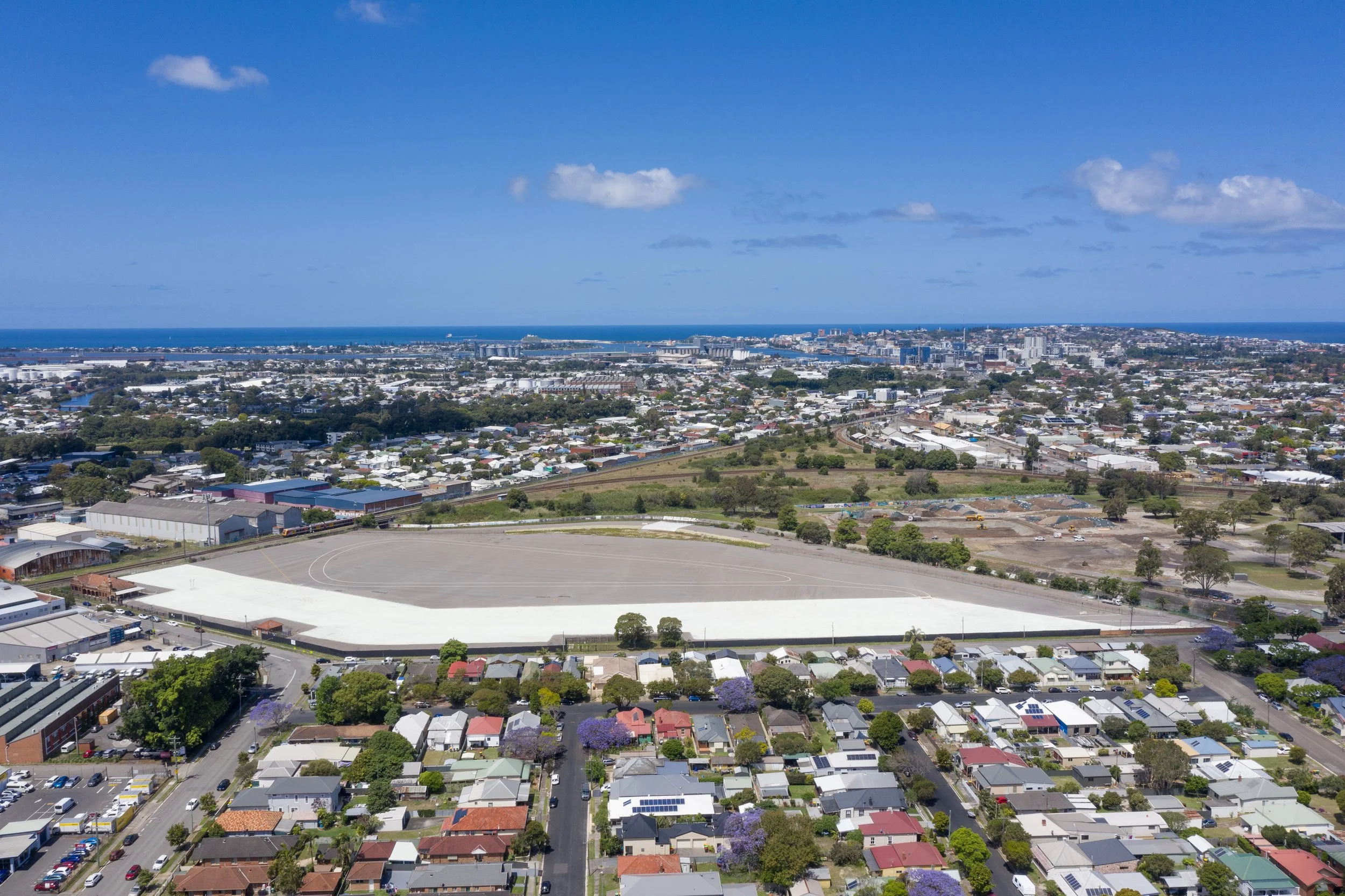Aerial view of a coastal city with residential and commercial buildings, green trees, a sports field, and the ocean in the background under a blue sky with scattered clouds.