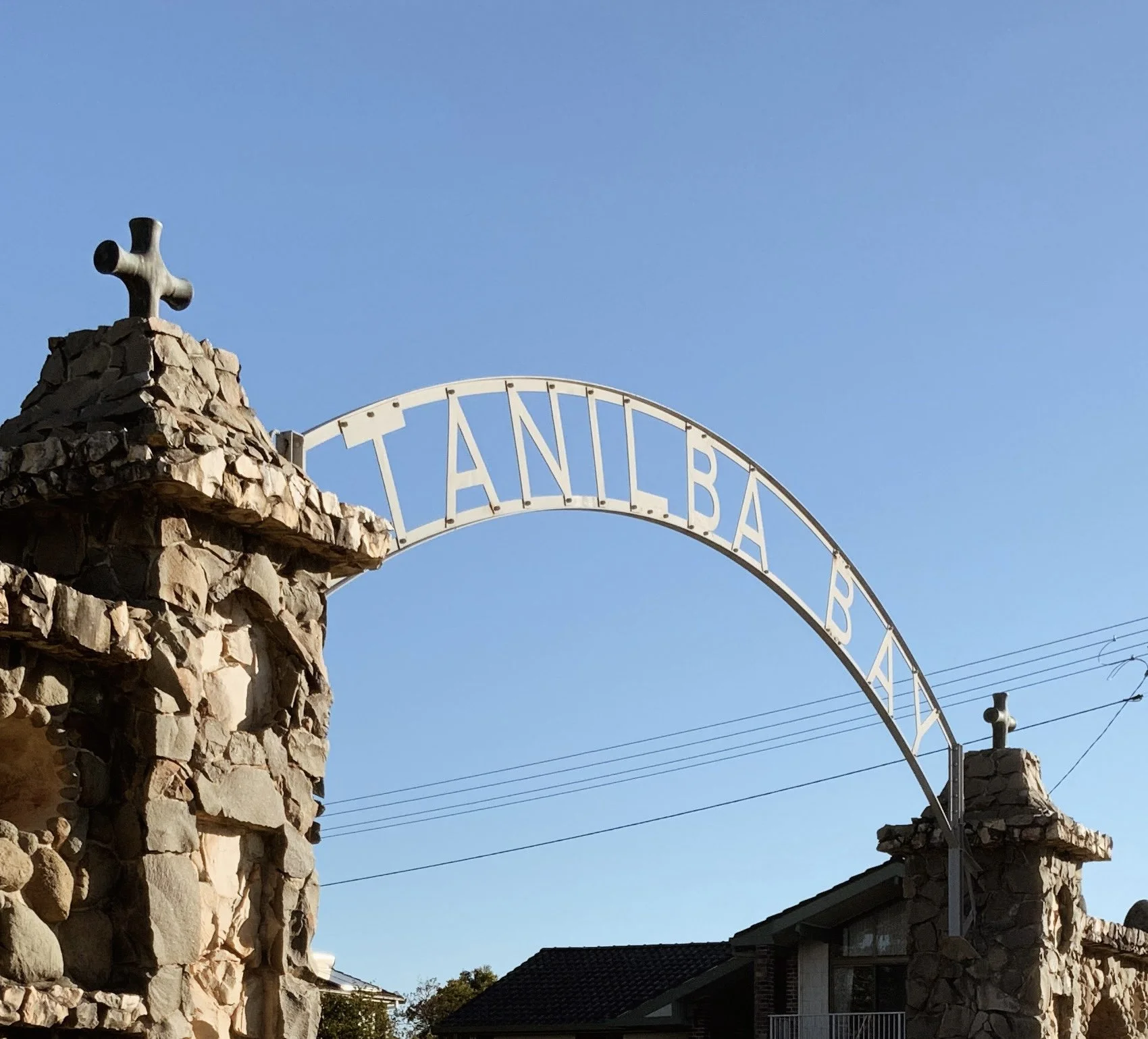Photo of a stone wall with a cross on top, and part of an arched sign that reads 'TAN' and 'BA'. The sky is clear and blue, with a power line in the background.