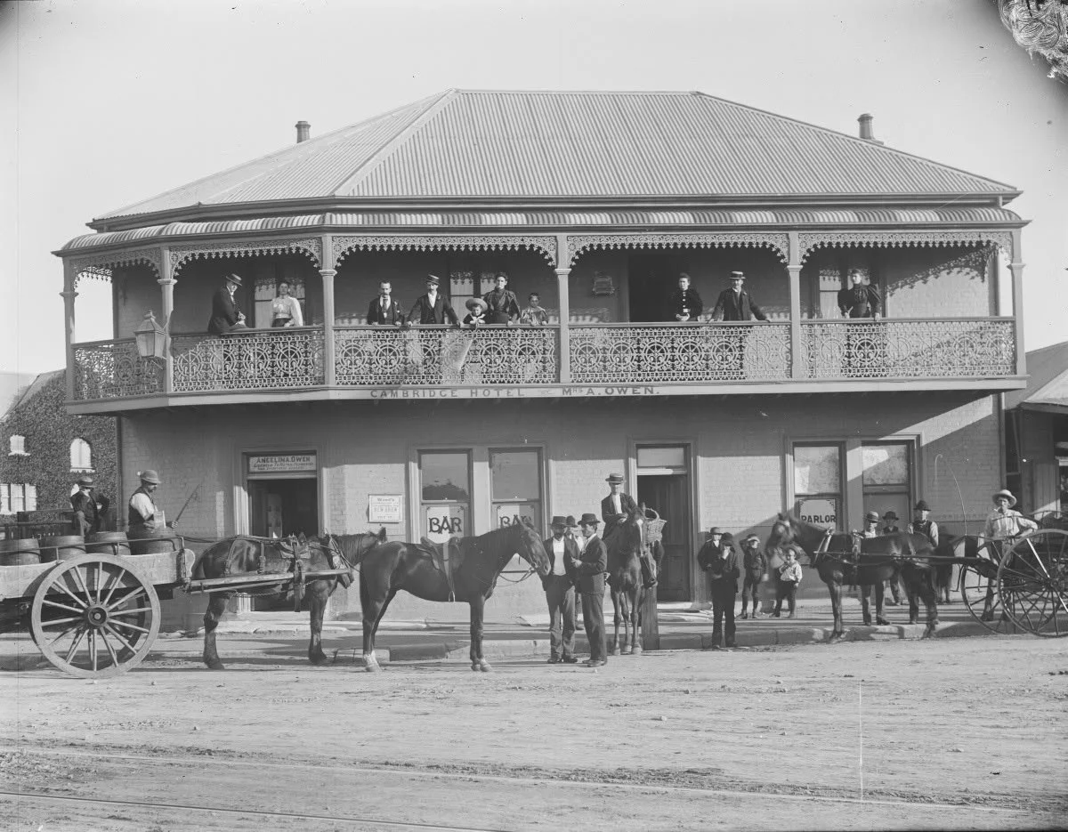 Black and white photo of a two-story building labeled 'Cambridge Hotel' with a sign for 'Mrs. A. Owen.' The upper balcony has seven people standing, dressed in early 20th-century clothing. The ground level has several people gathered outside, some on horseback and some standing on the sidewalk. There are horse-drawn carriages parked nearby and signs indicating a bar and parlor inside.