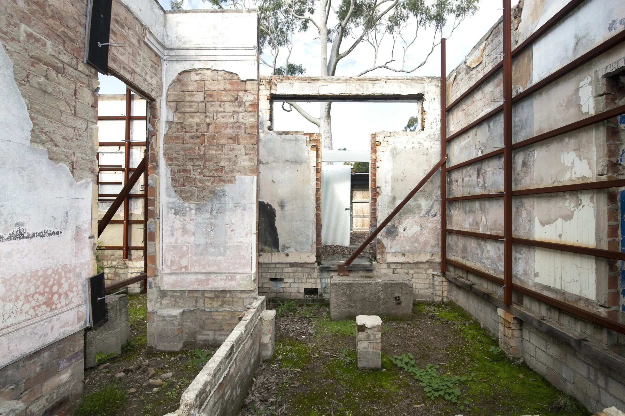 View of a construction site with partially built brick walls and wooden support beams inside a building, with a tree visible through the open roof area.