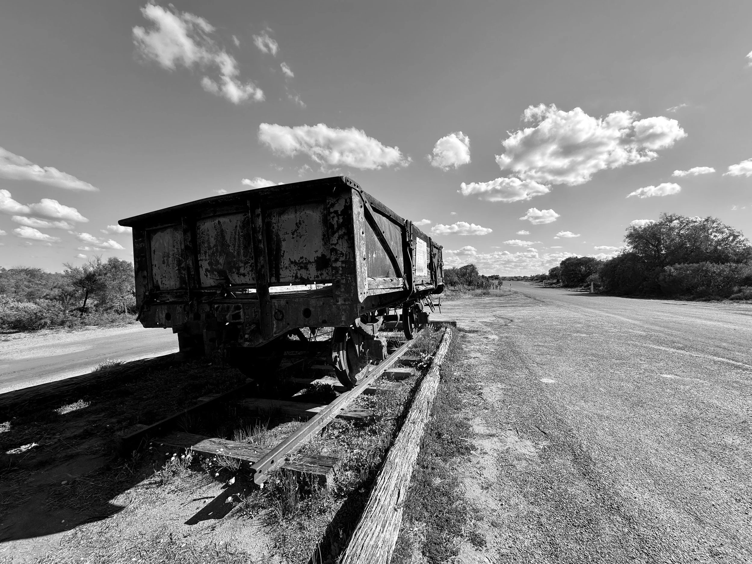 Black and white photo of an abandoned, weathered railroad freight car on a track beside a dirt road in a rural area with trees and a cloudy sky.