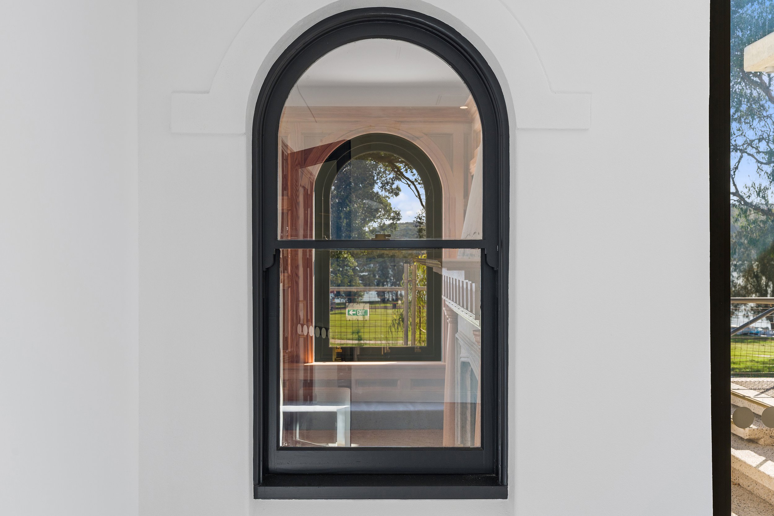 A multi-pane arched window with a black frame set in a white wall, reflecting trees and sky outside.