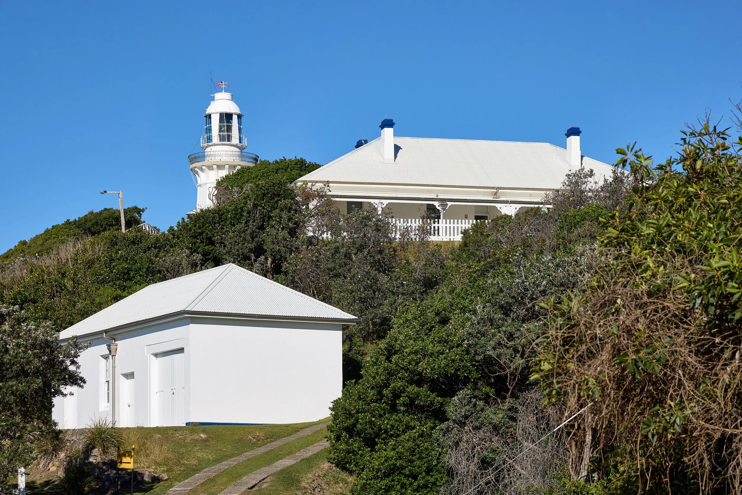 A white lighthouse with a small white building nearby, green bushes and trees surrounding it, and a clear blue sky overhead.