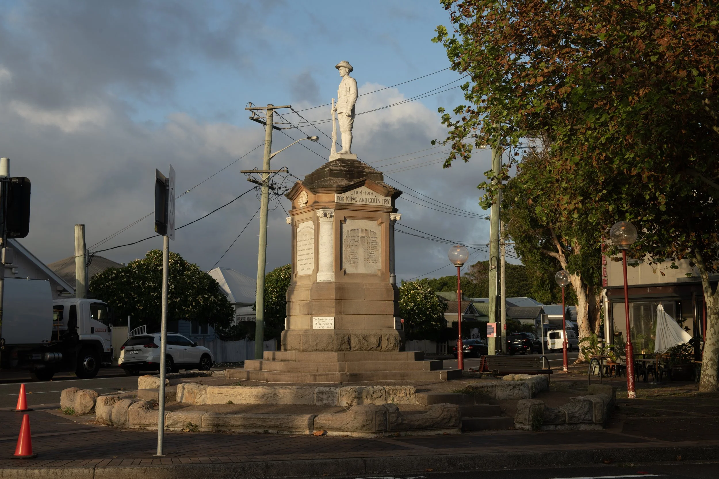 A war memorial with a statue of a soldier on top, surrounded by trees, parked cars, streetlights, and electrical poles, with a cloudy sky in the background.