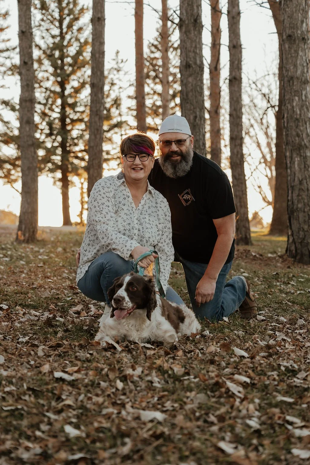 A smiling woman and a man kneeling in the woods with a brown and white spaniel dog, during sunset autumn, surrounded by tall trees and fallen leaves.