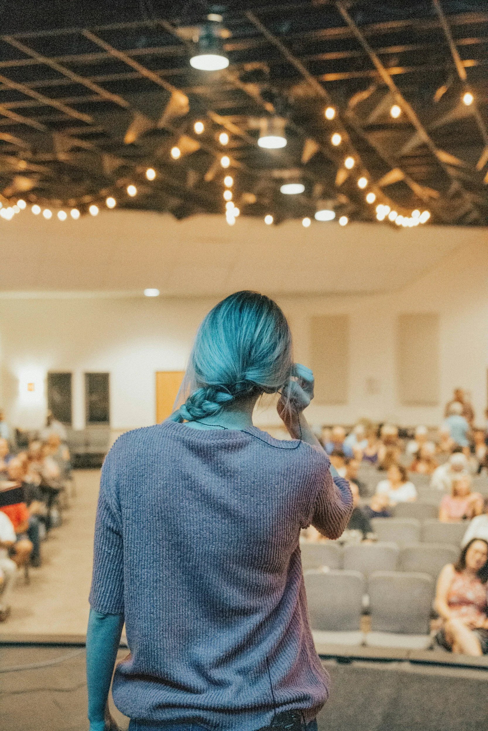 A person with blue hair and a gray sweater standing on a stage facing an audience in a large room with a high ceiling and string lights overhead.