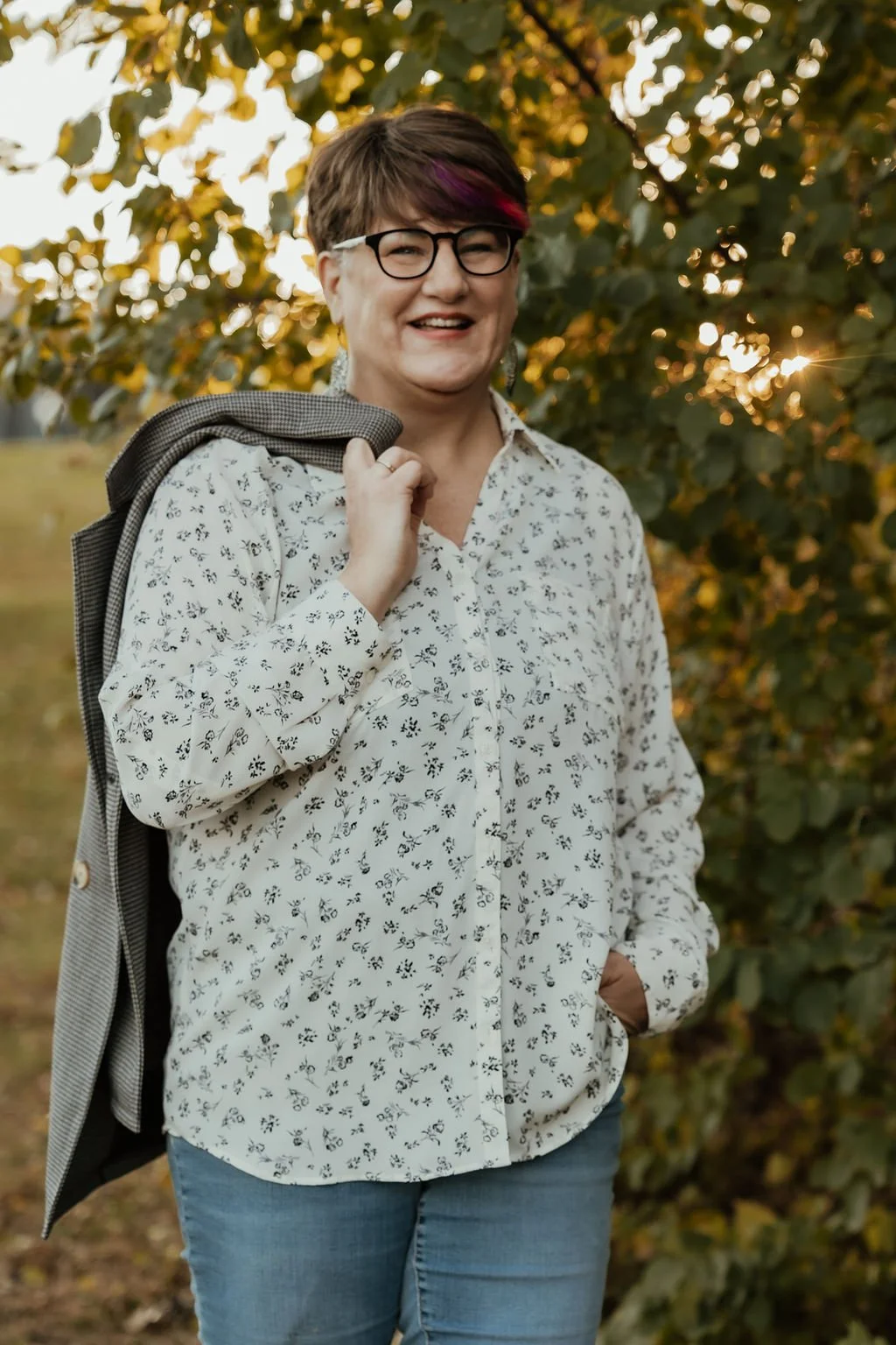 A smiling woman with short hair, glasses, and a white blouse with a small black floral pattern, stands outdoors in front of green foliage during sunset, holding a jacket over her shoulder.