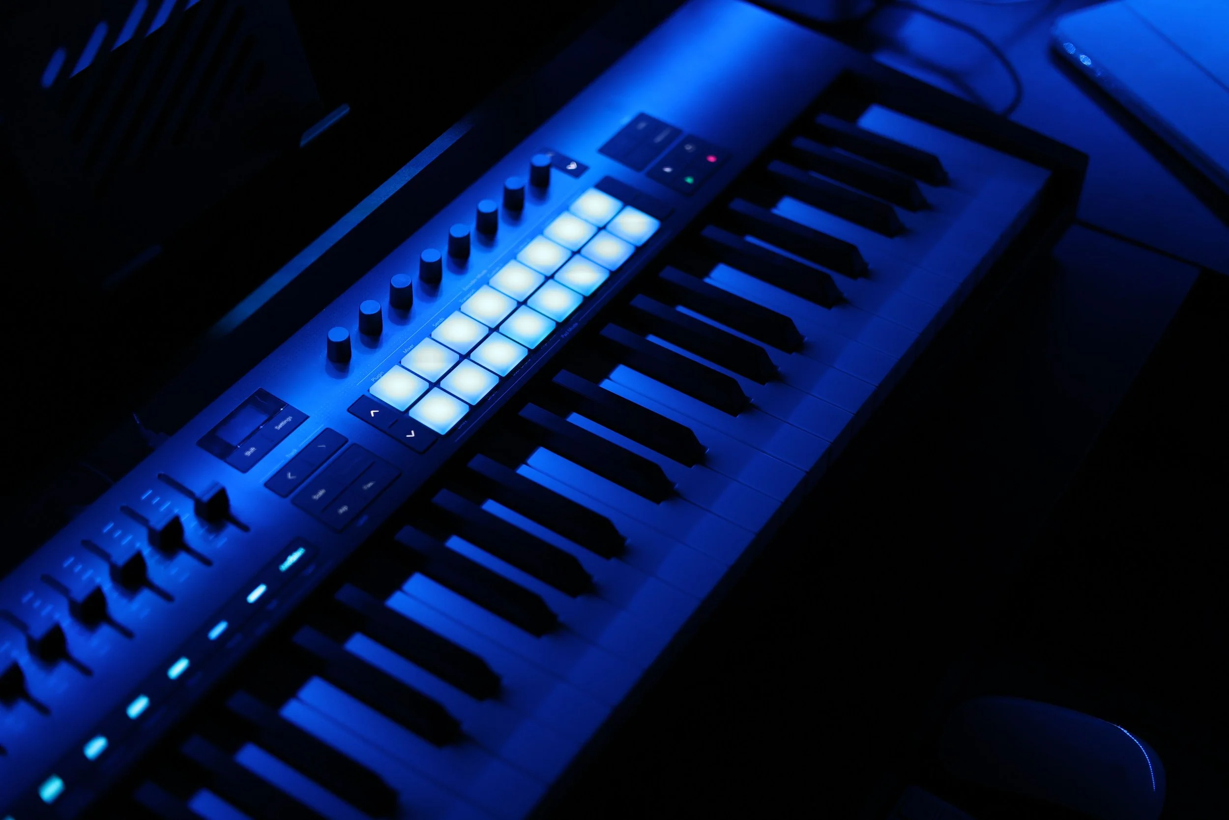 A digital music production keyboard with illuminated pads and knobs, lit with blue lighting in a dark environment.