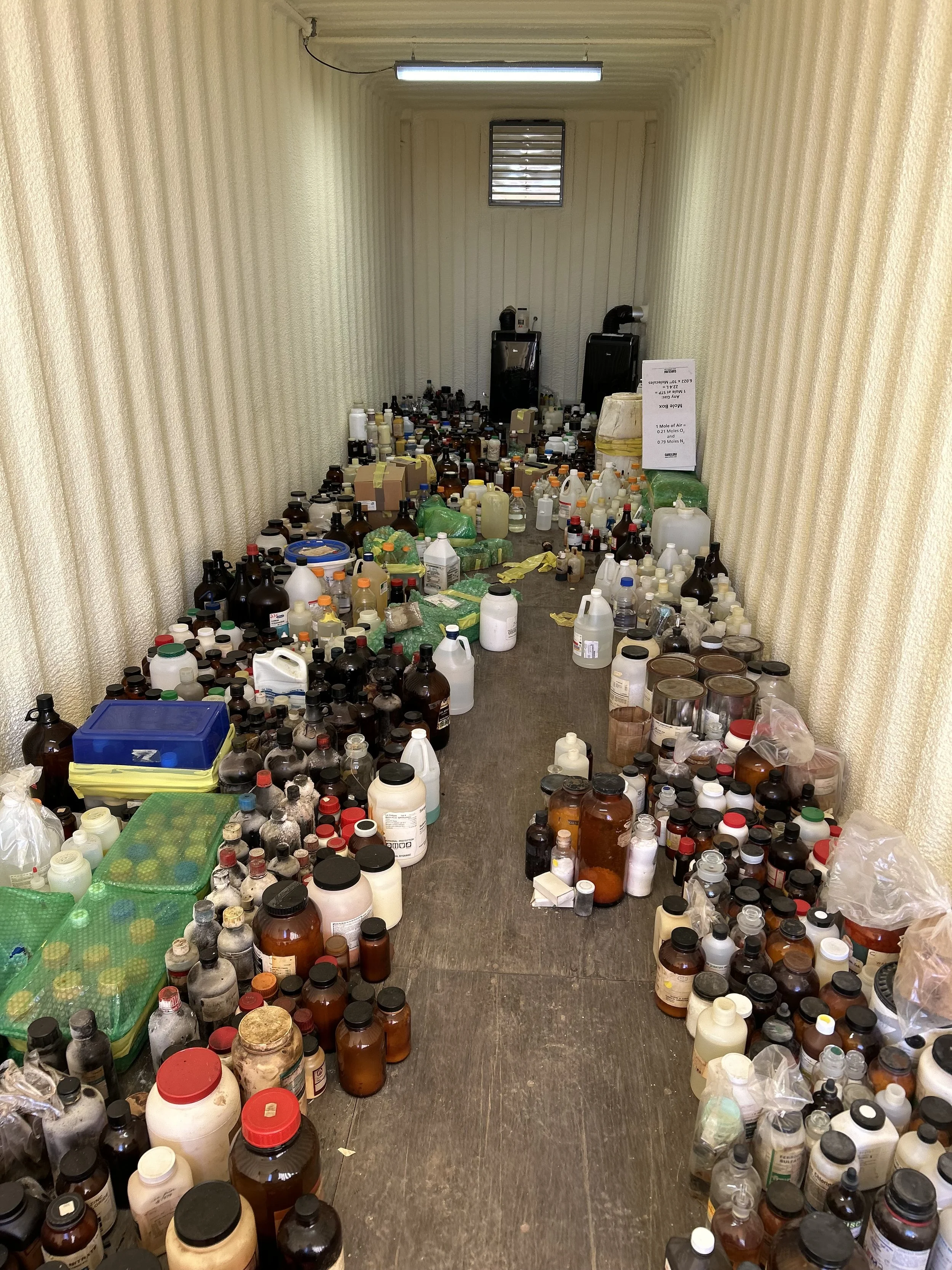 A room filled with numerous bottles and containers of various sizes and colors, some with labels. The bottles are scattered across the floor and stacked on each other. There are two large black machines or containers at the far end, and a small window and fluorescent light on the ceiling, with beige textured walls.