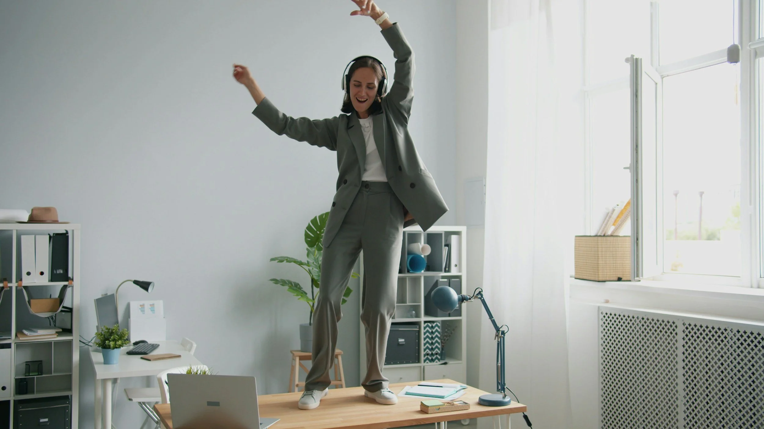 Une femme d'affaires en costume gris, portant un casque avec micro, danse sur une table dans un bureau lumineux avec des étagères, une plante verte, un ordinateur portable, un lampadaire et une fenêtre.