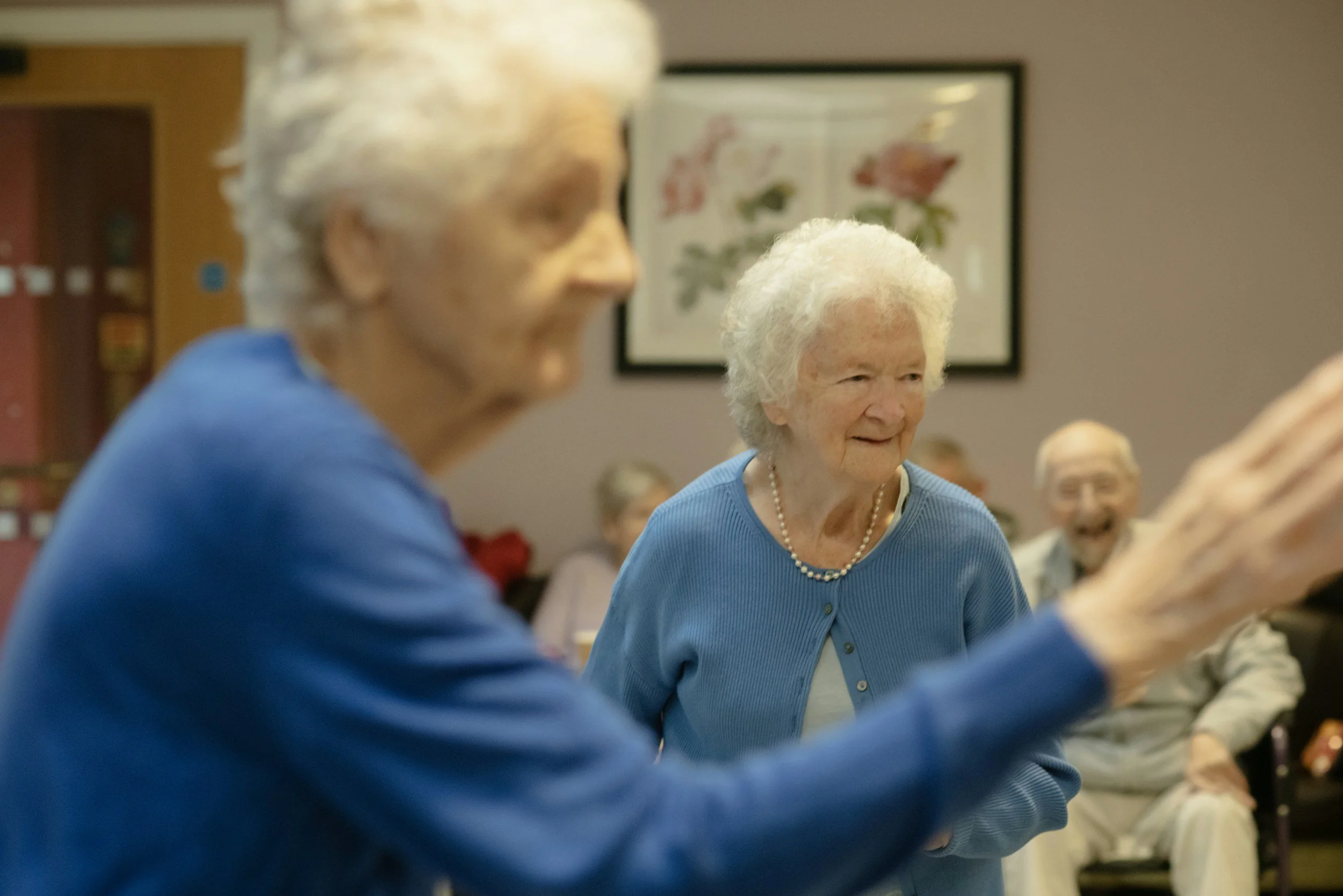 Deux dames âgées souriantes jouent à un jeu de société lors d'une réunion entre amis ou en famille dans une salle décorée avec des illustrations botaniques.
