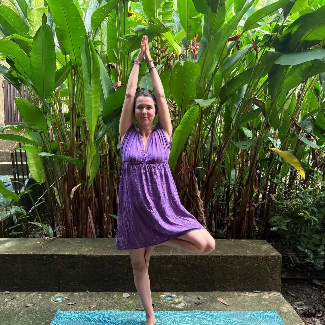 Une femme pratiquant le yoga en position de l'arbre, debout sur une jambe, les mains levées au-dessus de la tête, dans un jardin entouré de grandes feuilles vertes.